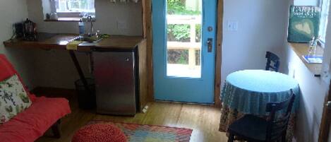 A compact kitchen with a refrigerator, a sink, and a window above the door. There's a dining area with a round table and a chair.