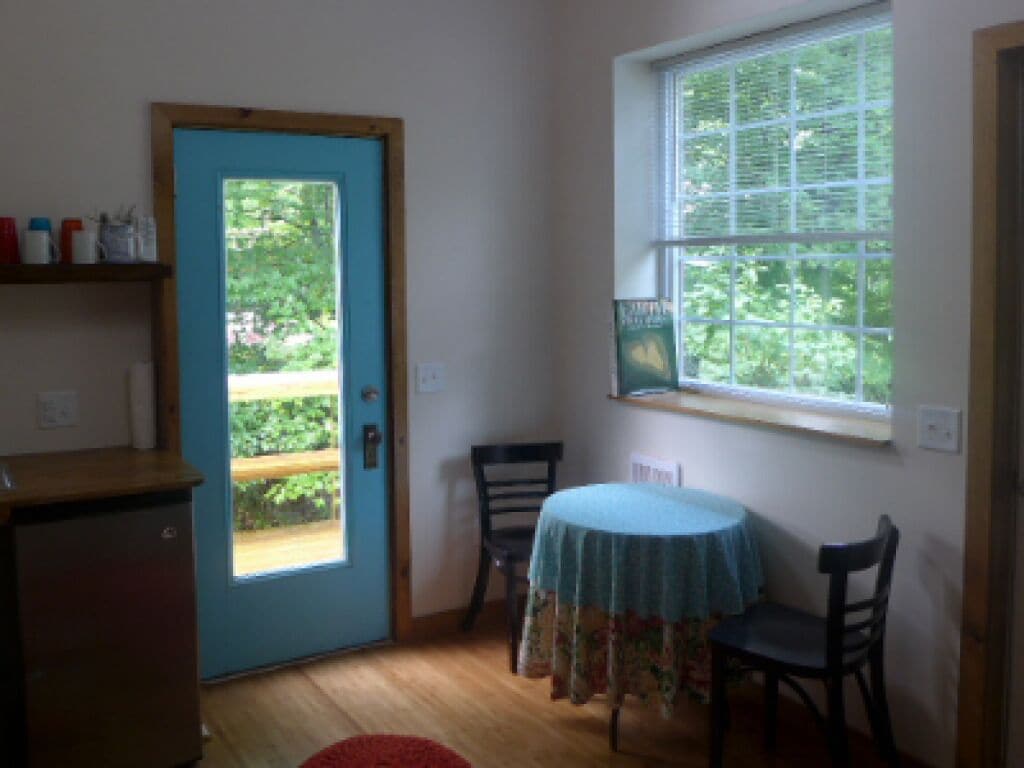 A room with a blue door, a small table with a floral tablecloth, two chairs, a window with blinds, and a wooden shelf with bottles.