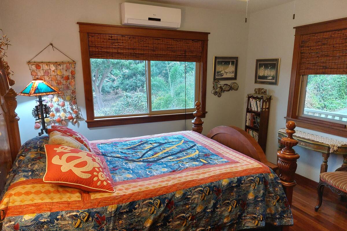 A bedroom with a large bed, a window with a view of greenery, a bookshelf, and a decorative lamp.
