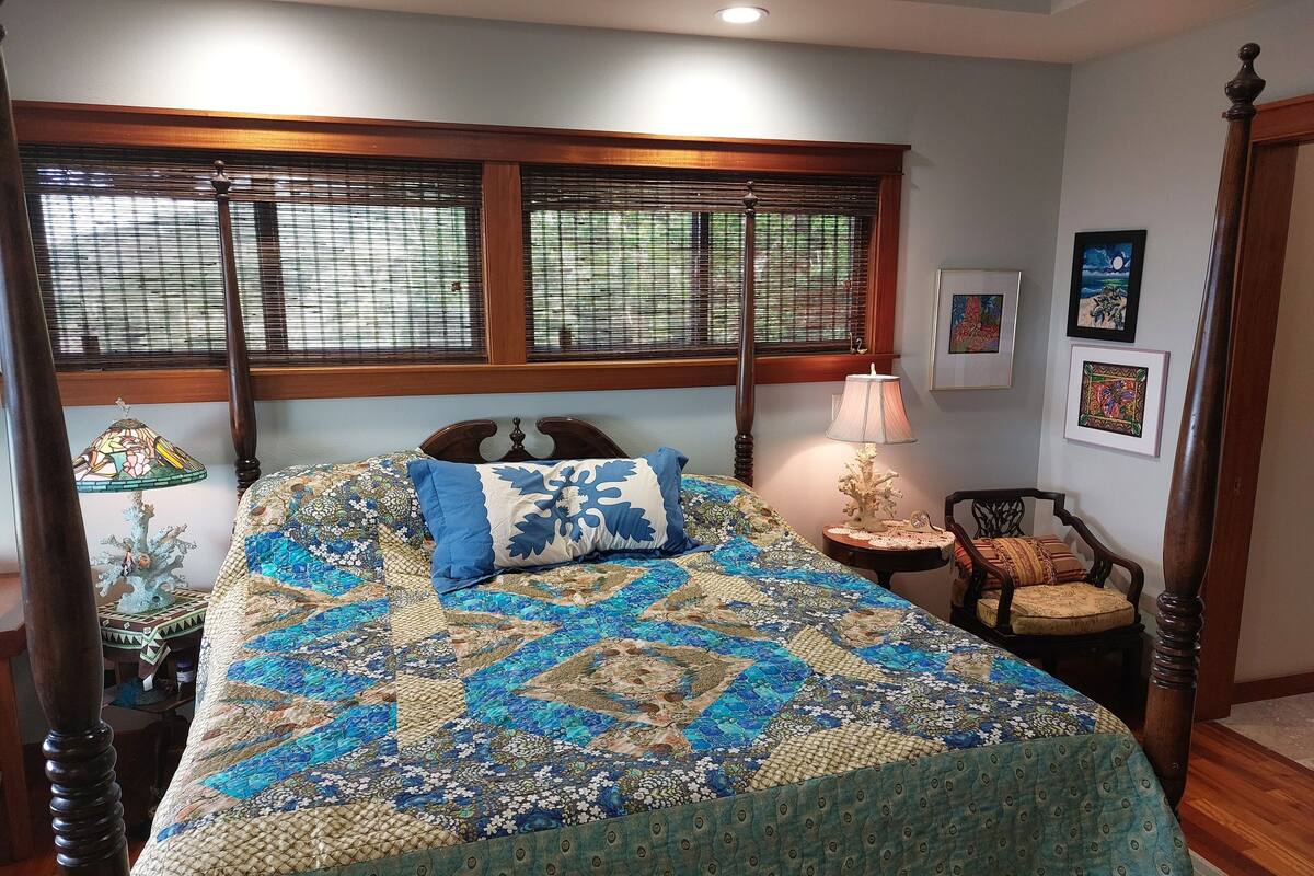 A four-poster bed with a quilt, a nightstand with a lamp, a chair, and framed artwork on the wall.