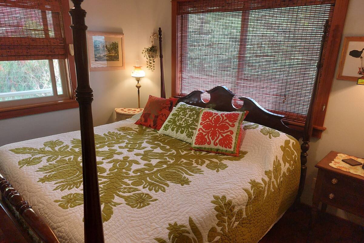 A four-poster bed with a floral bedspread, a nightstand with a lamp, and a window with blinds.
