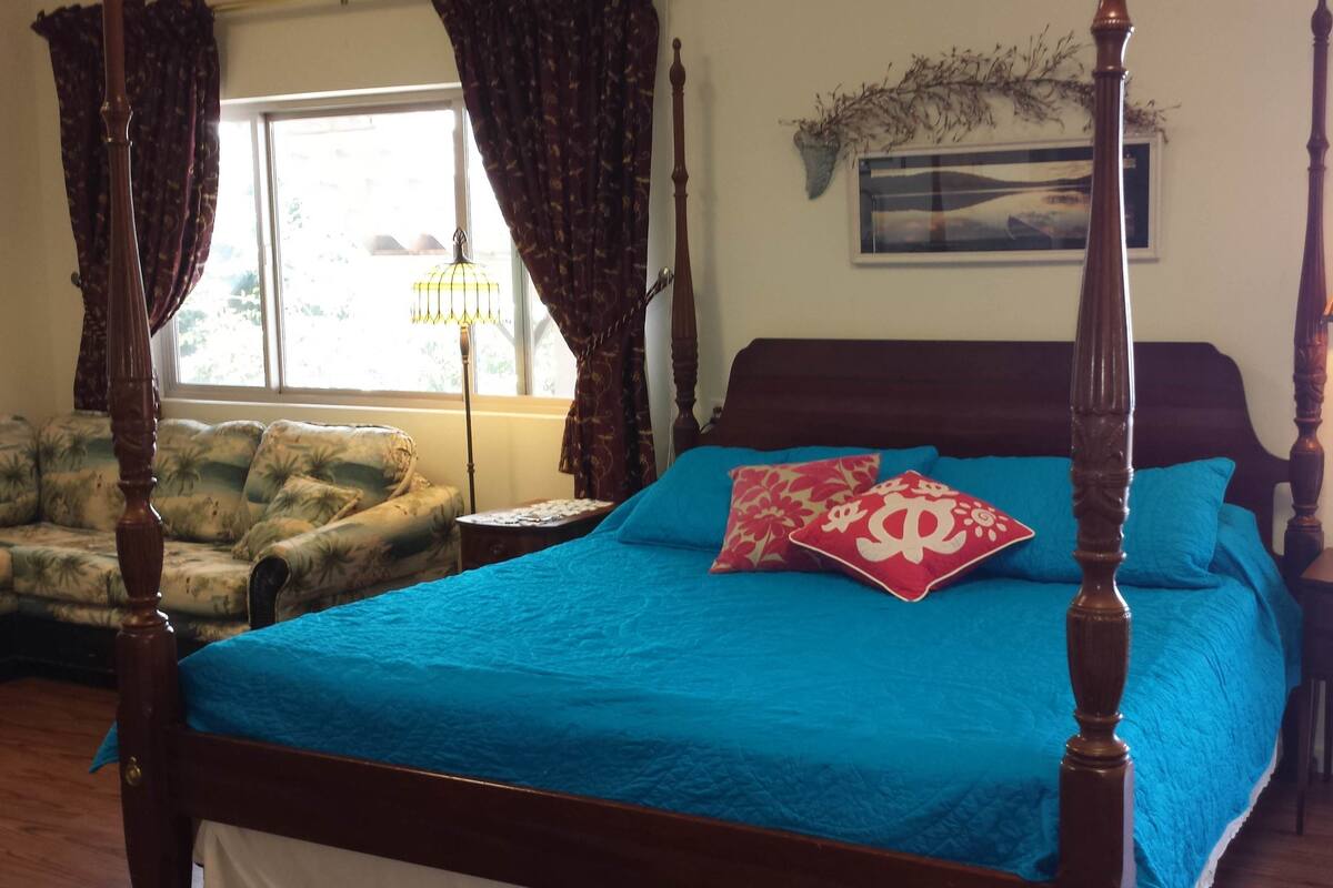 A four-poster bed with a blue bedspread and two decorative pillows, a wooden nightstand with a lamp, a floral-patterned armchair, and a window with curtains.