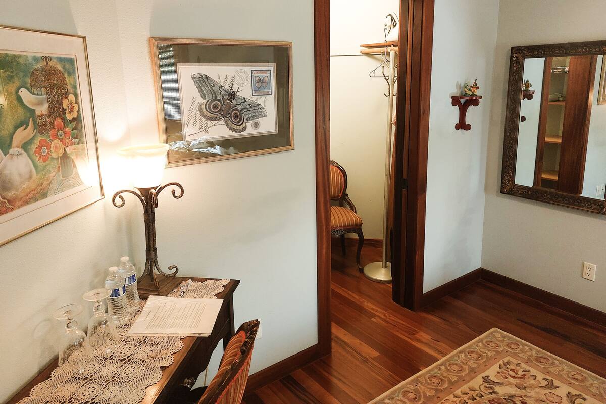 A room with a wooden floor, a dining table with chairs, a framed butterfly artwork, a mirror, and a door leading to another room.