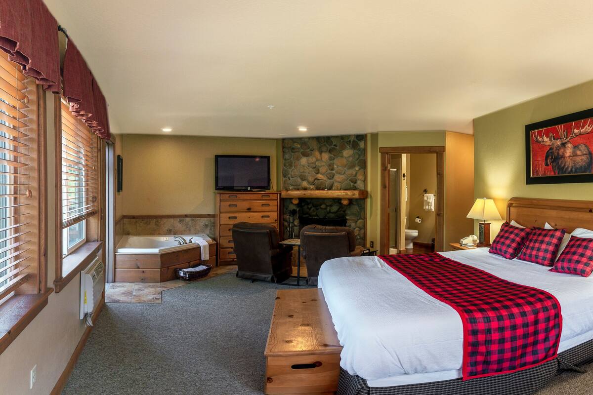A hotel room with a large bed, a wooden dresser, a stone fireplace, a television, and a bathroom visible through an open door.