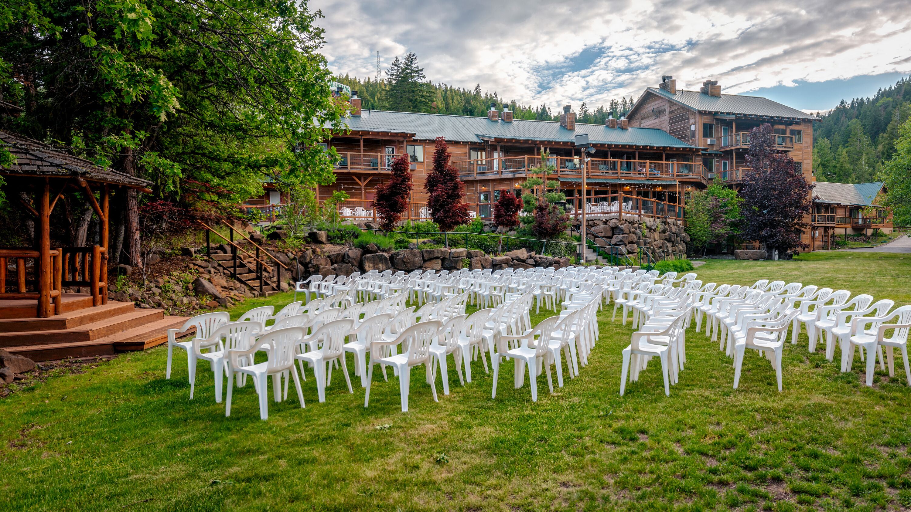 A lodge with a circular outdoor seating area arranged in rows of white plastic chairs, surrounded by a well-maintained lawn and a stone retaining wall.