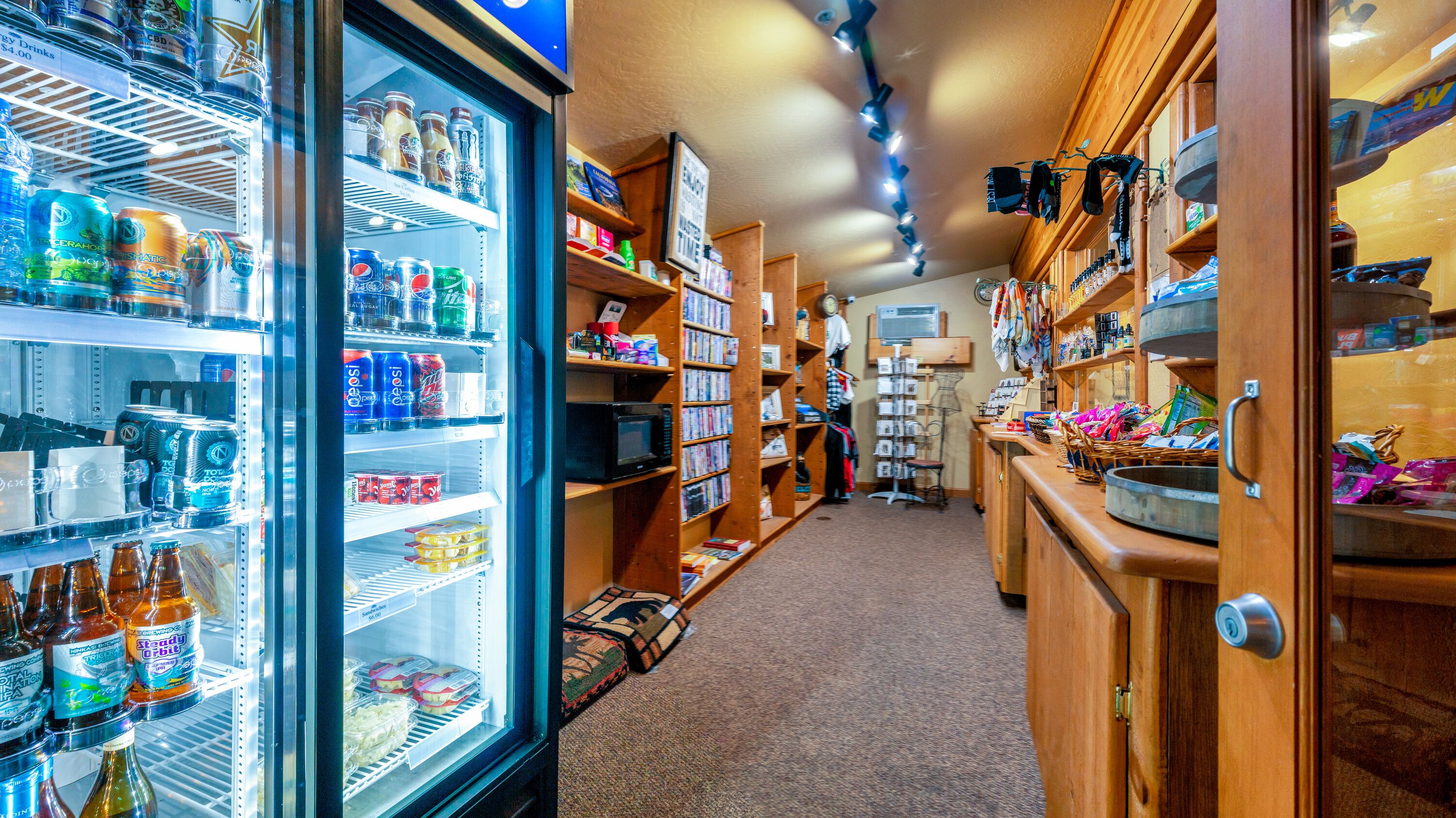 A store with shelves stocked with various beverages and snacks, a microwave, and a display of hats and other items.