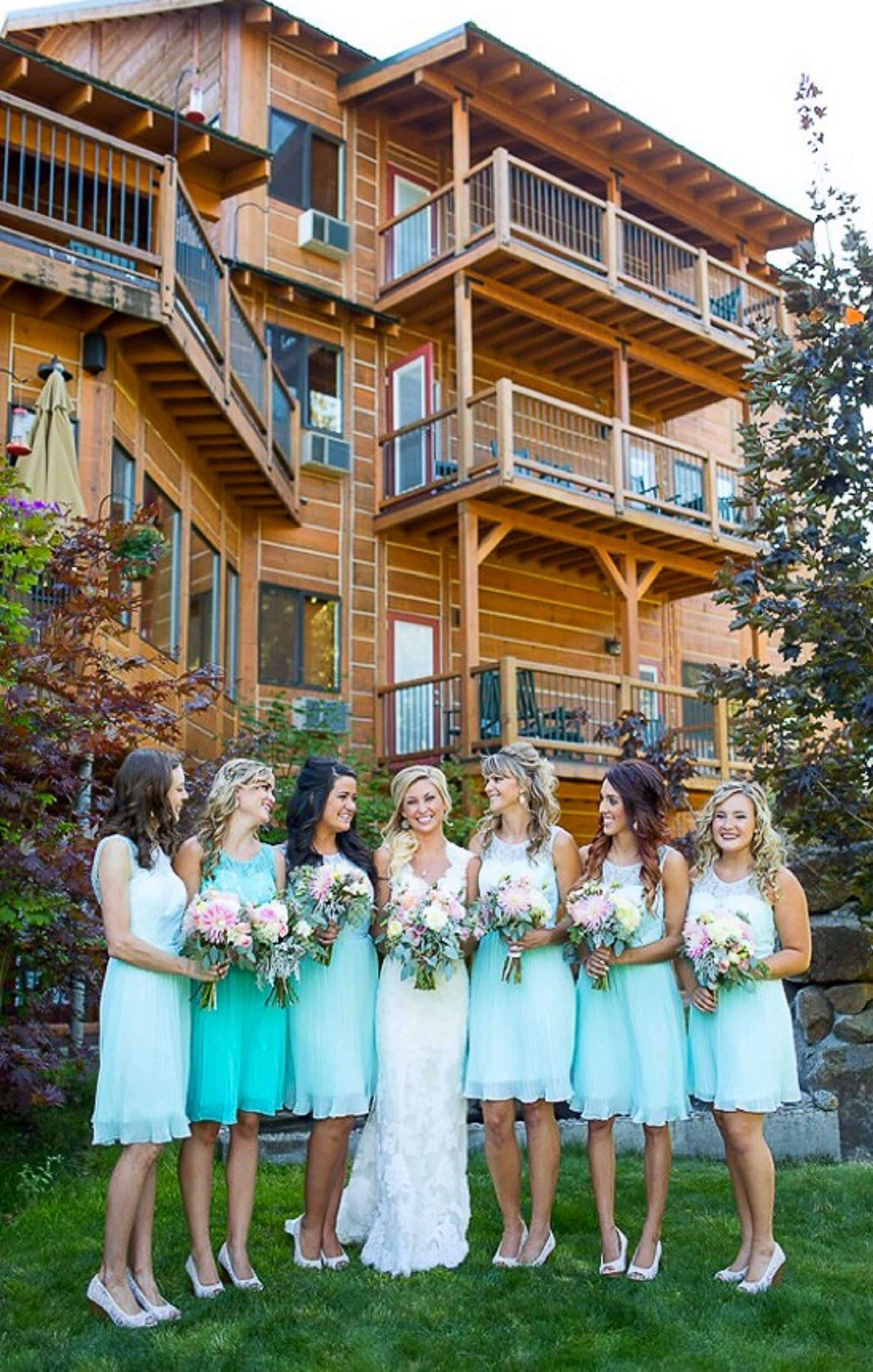Un grupo de mujeres con vestidos azul claro y una novia con vestido blanco, de pie frente a un edificio de madera con balcones.