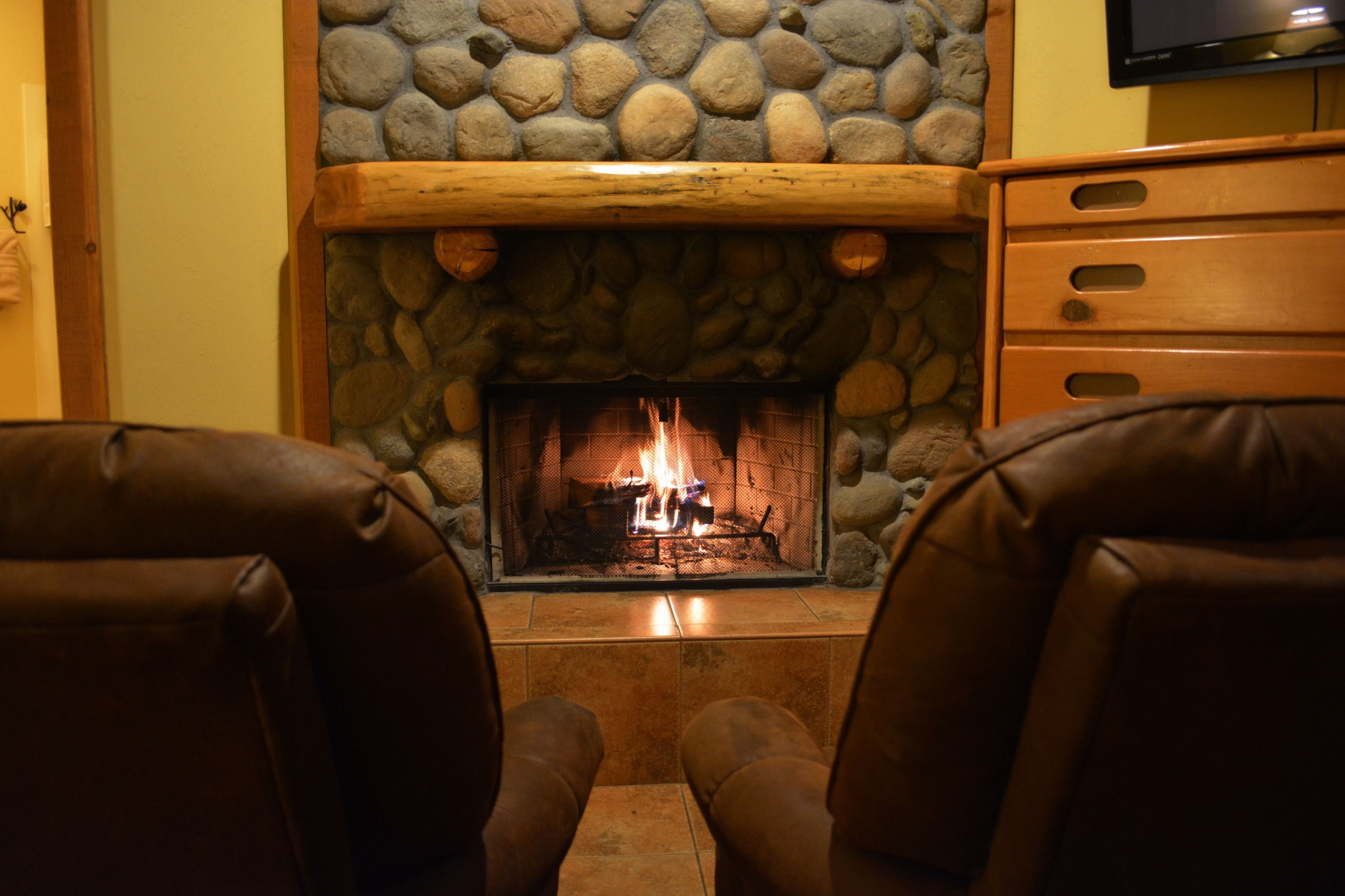 A stone fireplace with a wooden mantel, flanked by leather chairs, in a room with a wooden dresser and a television mounted on the wall.