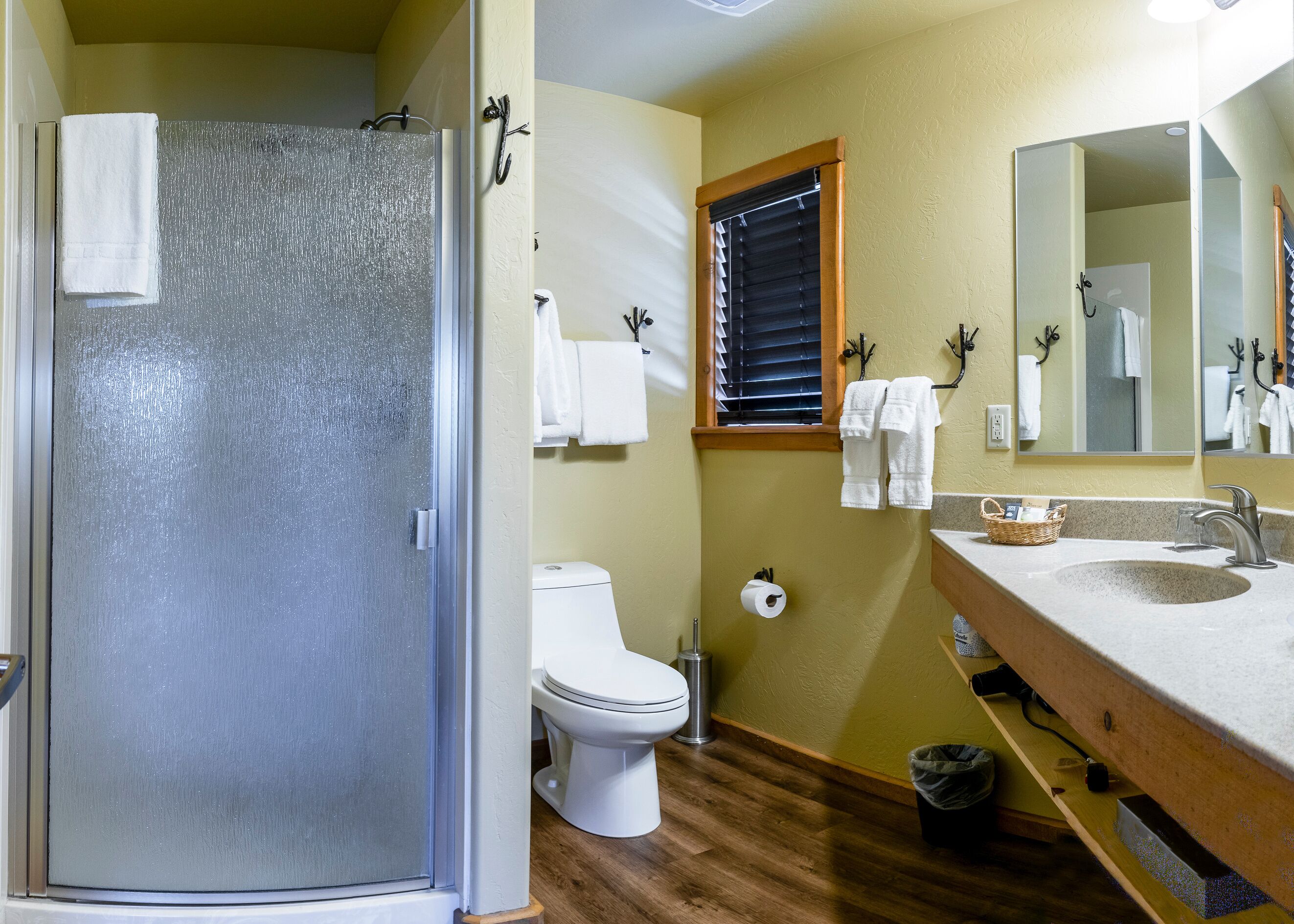 Un baño con mampara de vidrio, inodoro, lavamanos con espejo y toallas colgadas en ganchos.