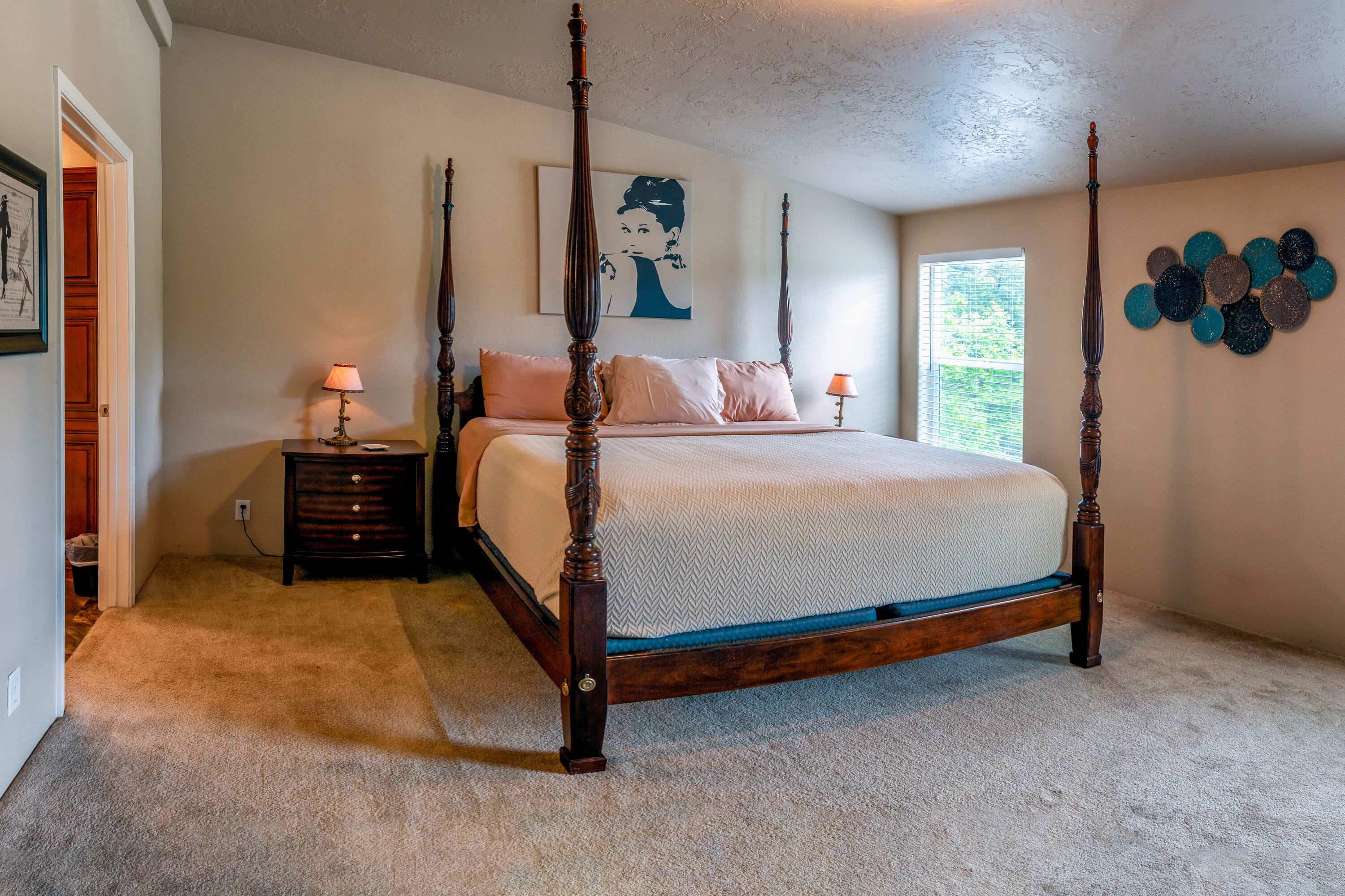 A four-poster bed with a wooden frame, a bedside table with a lamp, a wall-mounted artwork, and a window with blinds.