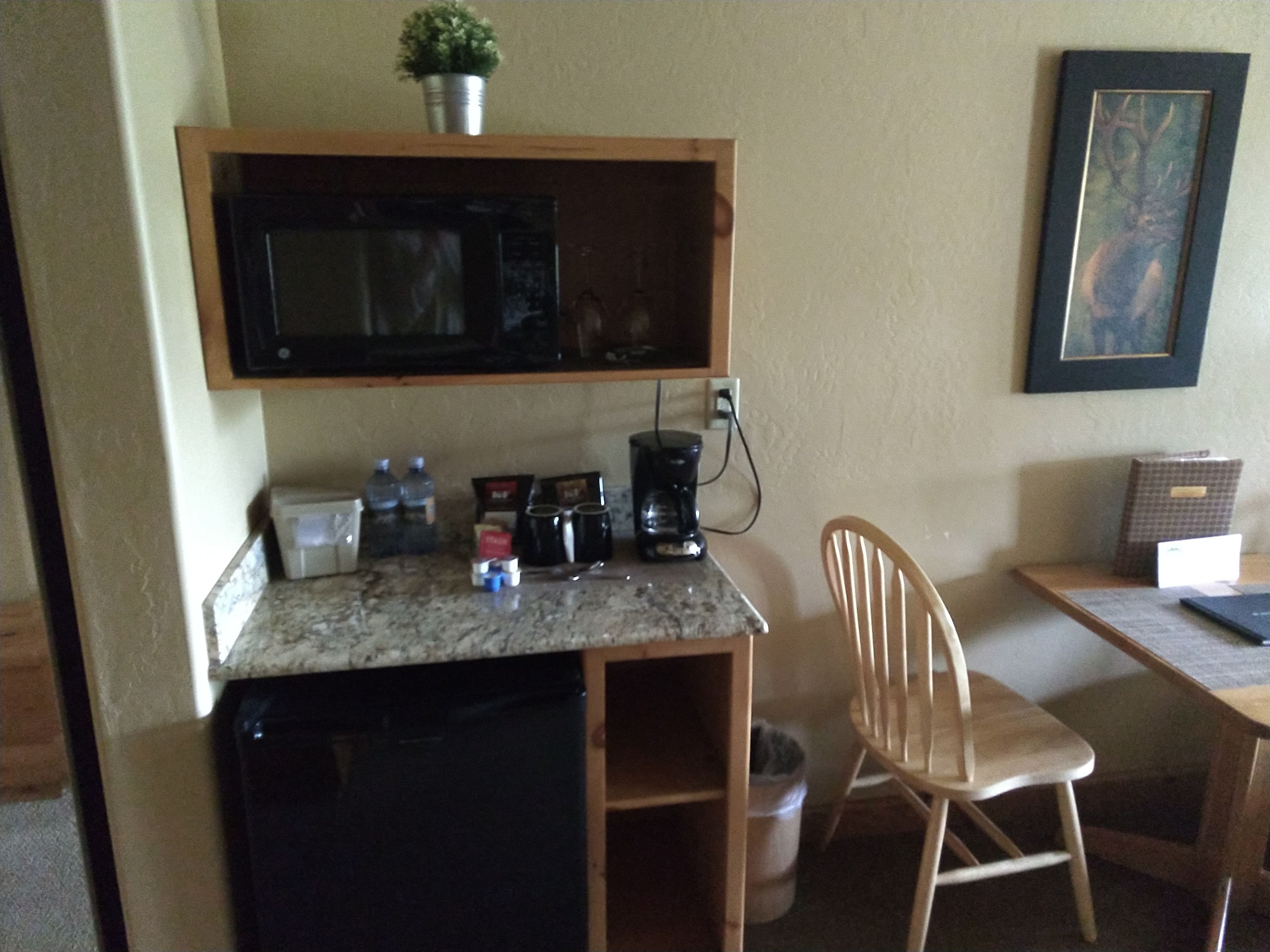 A compact kitchenette with a microwave, coffee maker, and storage containers. A chair is positioned near the counter, and a trash can is visible.