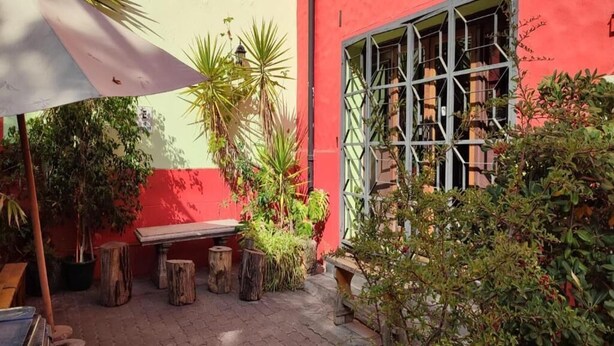 Un patio al aire libre con pared roja, bancos de madera, plantas en macetas y una ventana con celosía metálica.