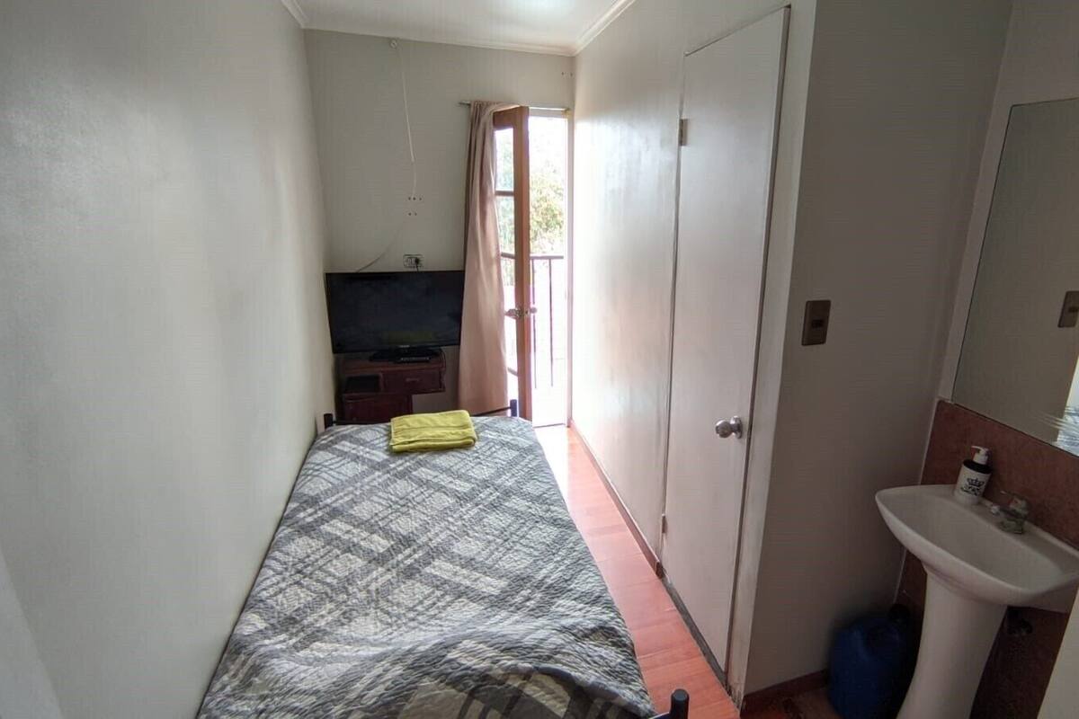 Ein schmaler Raum mit einem Bett, einem kleinen Schreibtisch, einem Fernseher, einer Tür und einem Badezimmer mit Waschbecken und Spiegel.