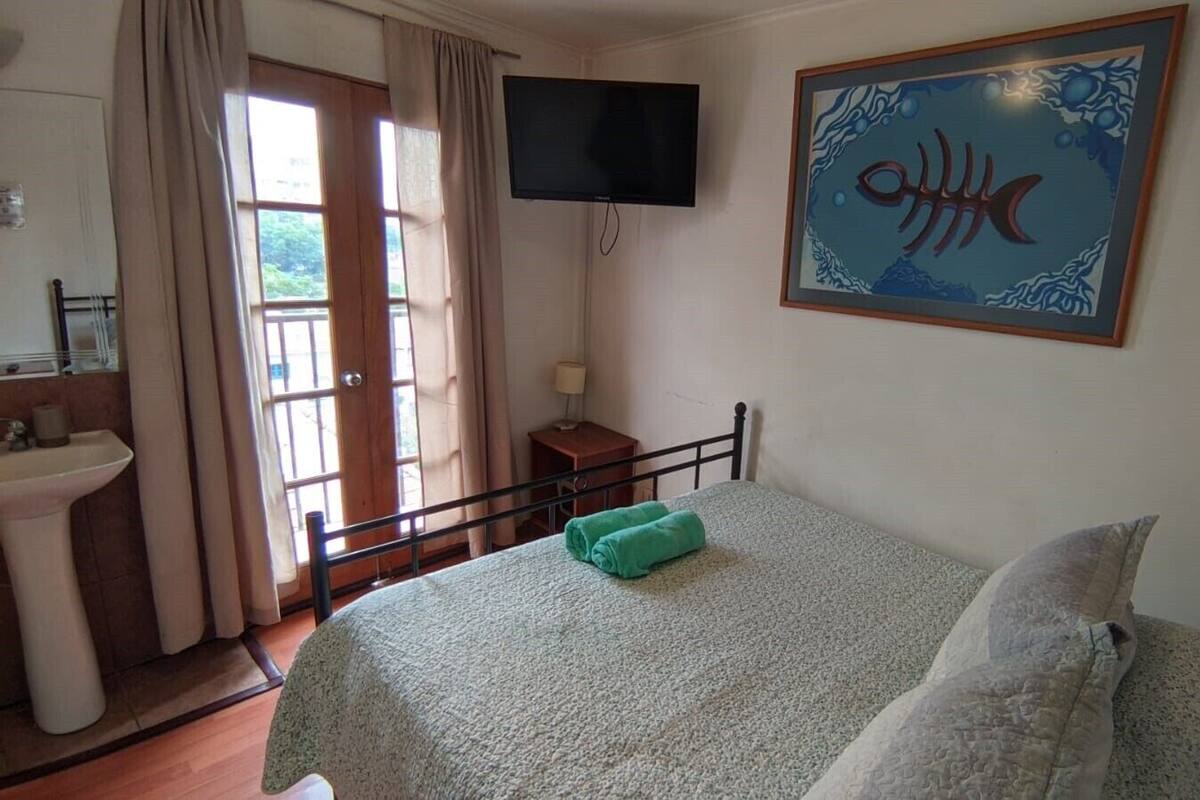 Ein Schlafzimmer mit einem Bett, einem Fernseher, einem Gemälde, einem Fenster mit Vorhängen und einem Waschbecken.