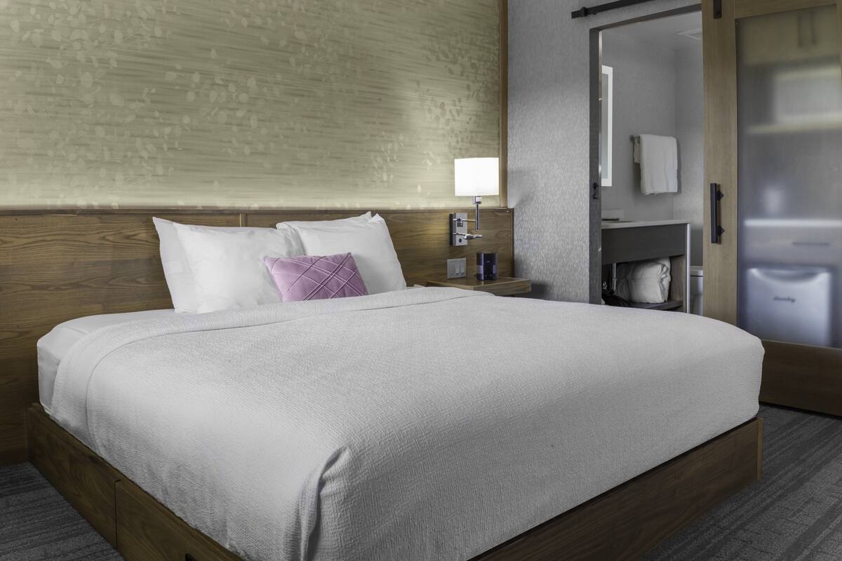 Una cama bien tendida con sábanas blancas y un cojín morado, una mesita de noche con lámpara, y un cabecero de madera.