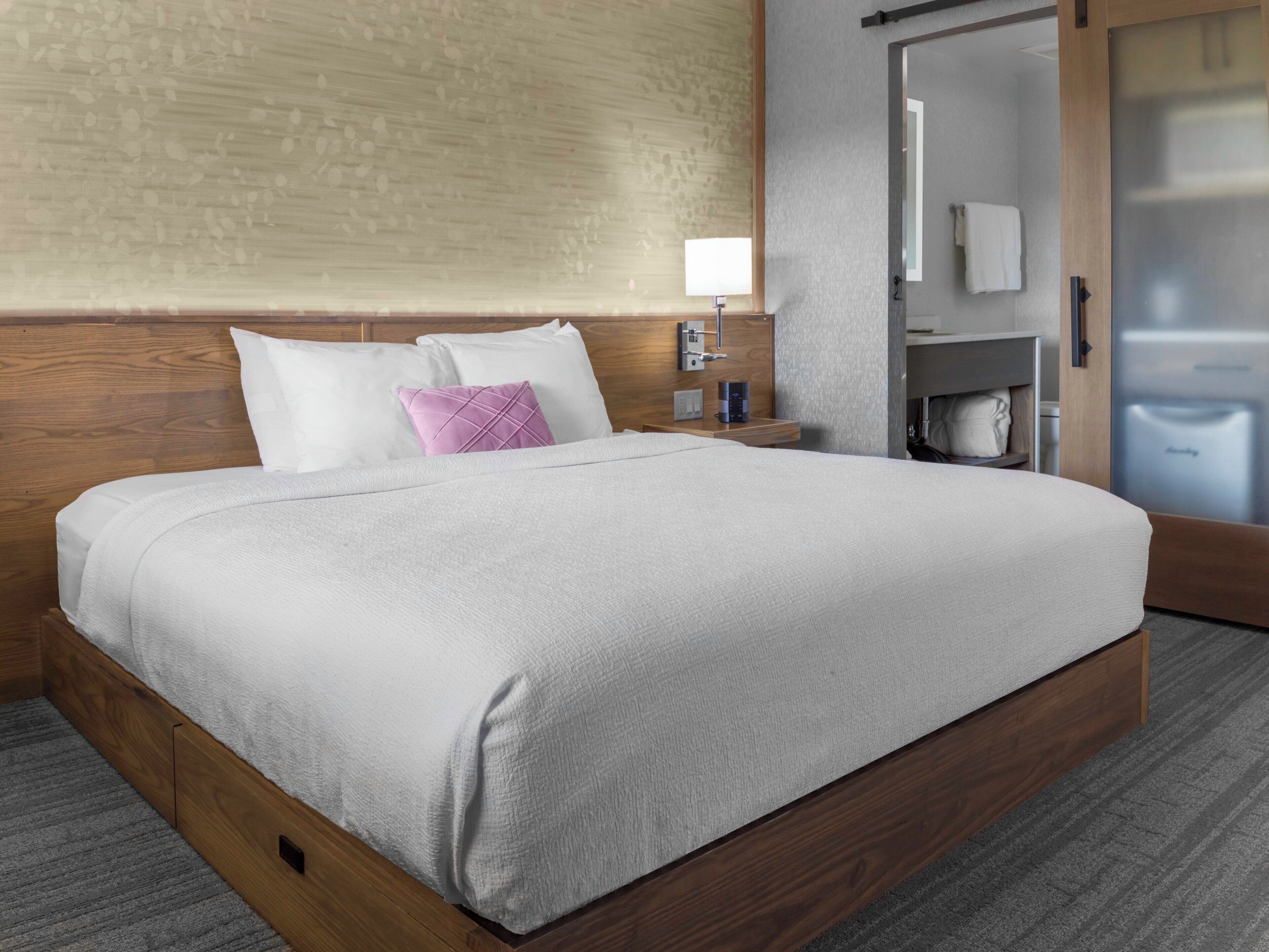 Cama bem arrumada com lençóis brancos e um travesseiro roxo, mesa de cabeceira com abajur e cabeceira de madeira.