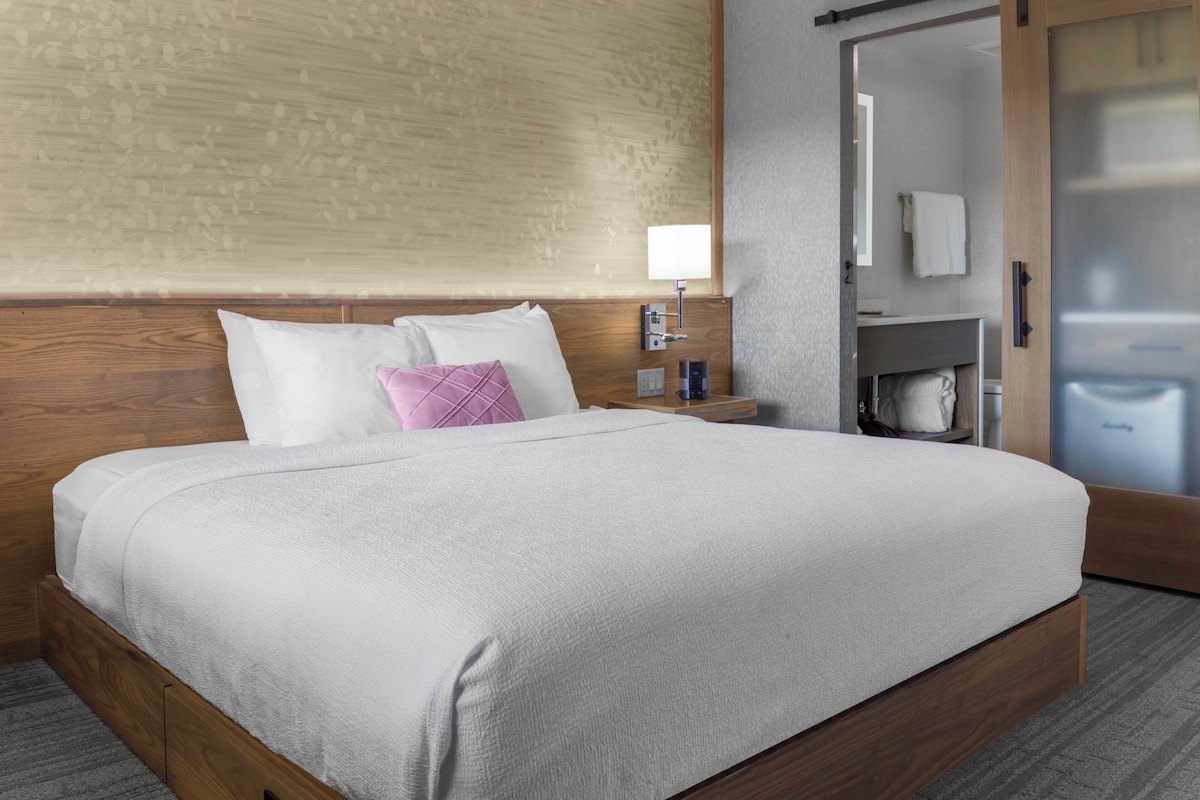 A neatly made bed with white linens and a purple pillow, a bedside table with a lamp, and a wooden headboard.