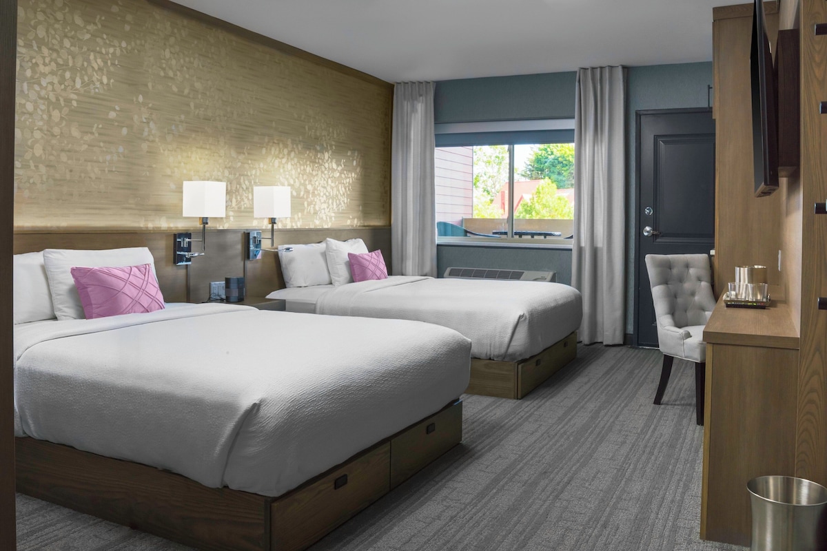 A hotel room with two beds, a chair, a desk, a trash can, and a window with curtains.