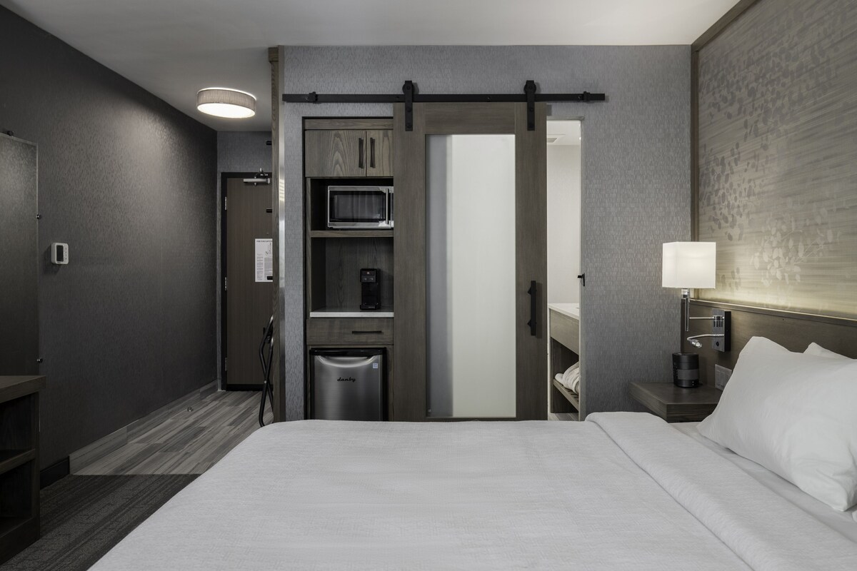 Ein Hotelzimmer mit einem Bett, einem Nachttisch mit Lampe, einer Mikrowelle, einem Kühlschrank, einem Schiebetürenschrank und einem kleinen Waschbecken.