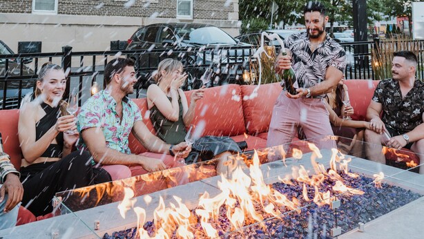 A group of people sitting on a red couch around a glass-top fire pit with flames.