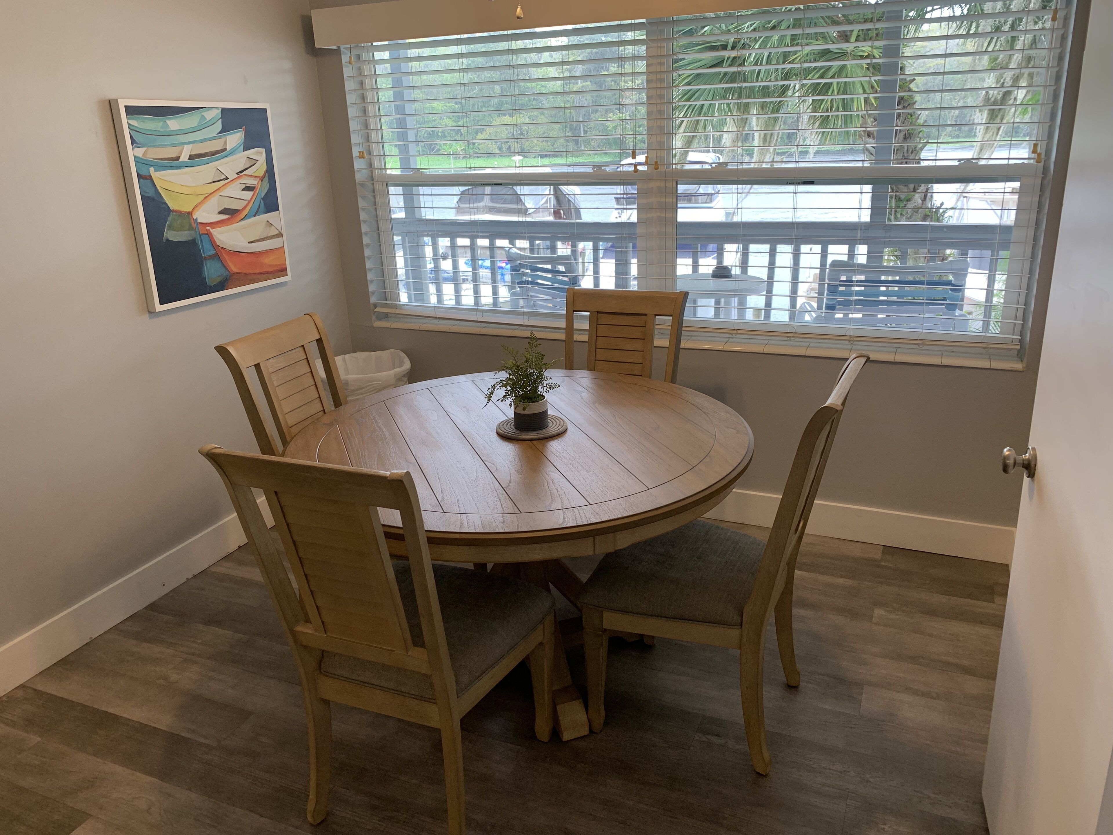 A dining area with a round wooden table, four chairs, a potted plant, and a window with a view of a deck and greenery.