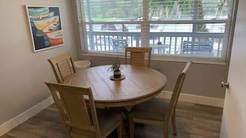 A dining area with a round wooden table, four chairs, a potted plant, and a window with a view of a deck and greenery.