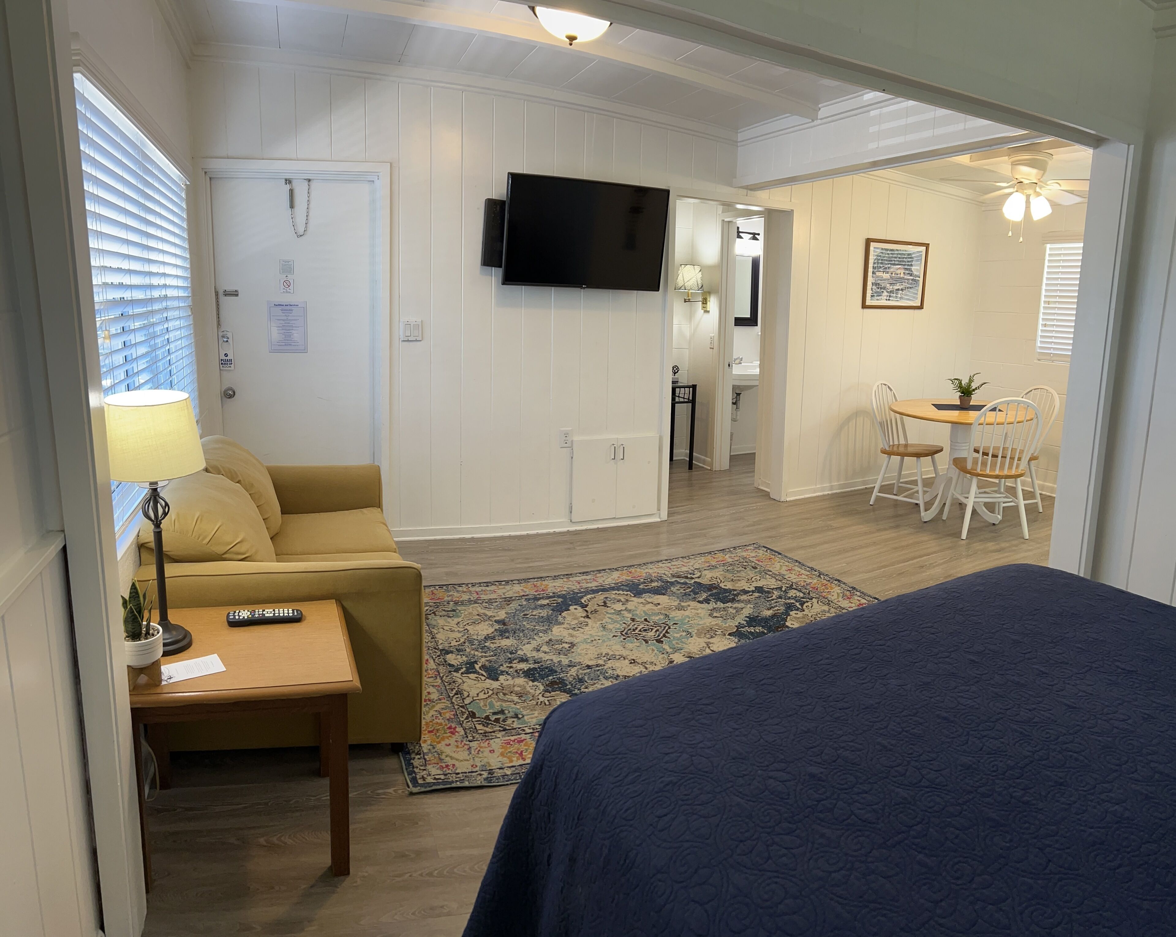 A room with a sofa, a small table with a lamp, a TV mounted on the wall, a bed, a dining area with a table and chairs, and a ceiling fan.