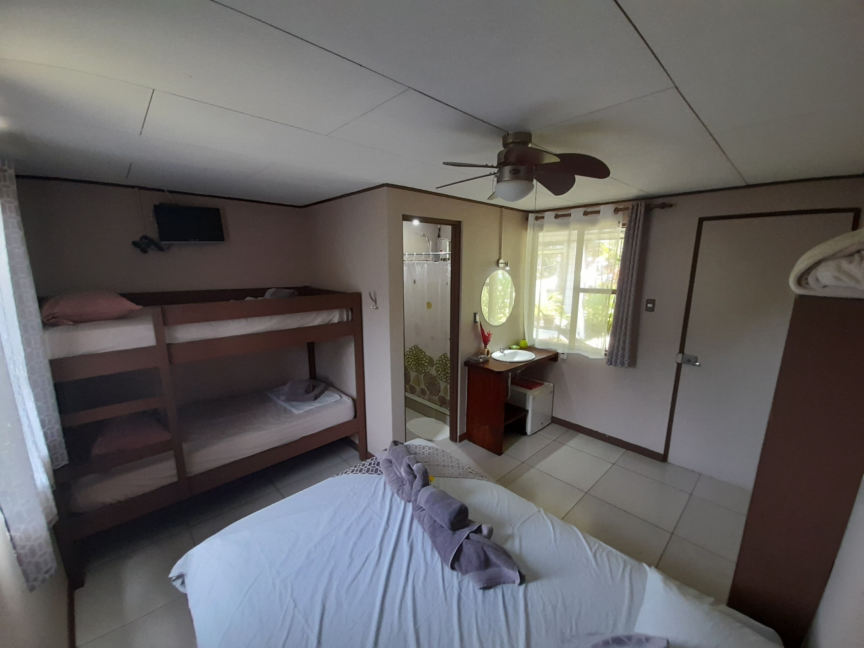 Une chambre avec des lits superposés, un lit simple, un ventilateur de plafond, une fenêtre avec des rideaux, une salle de bain avec un lavabo et un miroir, et une porte.