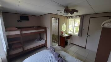 Une chambre avec des lits superposés, un lit simple, un ventilateur de plafond, une fenêtre avec des rideaux, une salle de bain avec un lavabo et un miroir, et une porte.