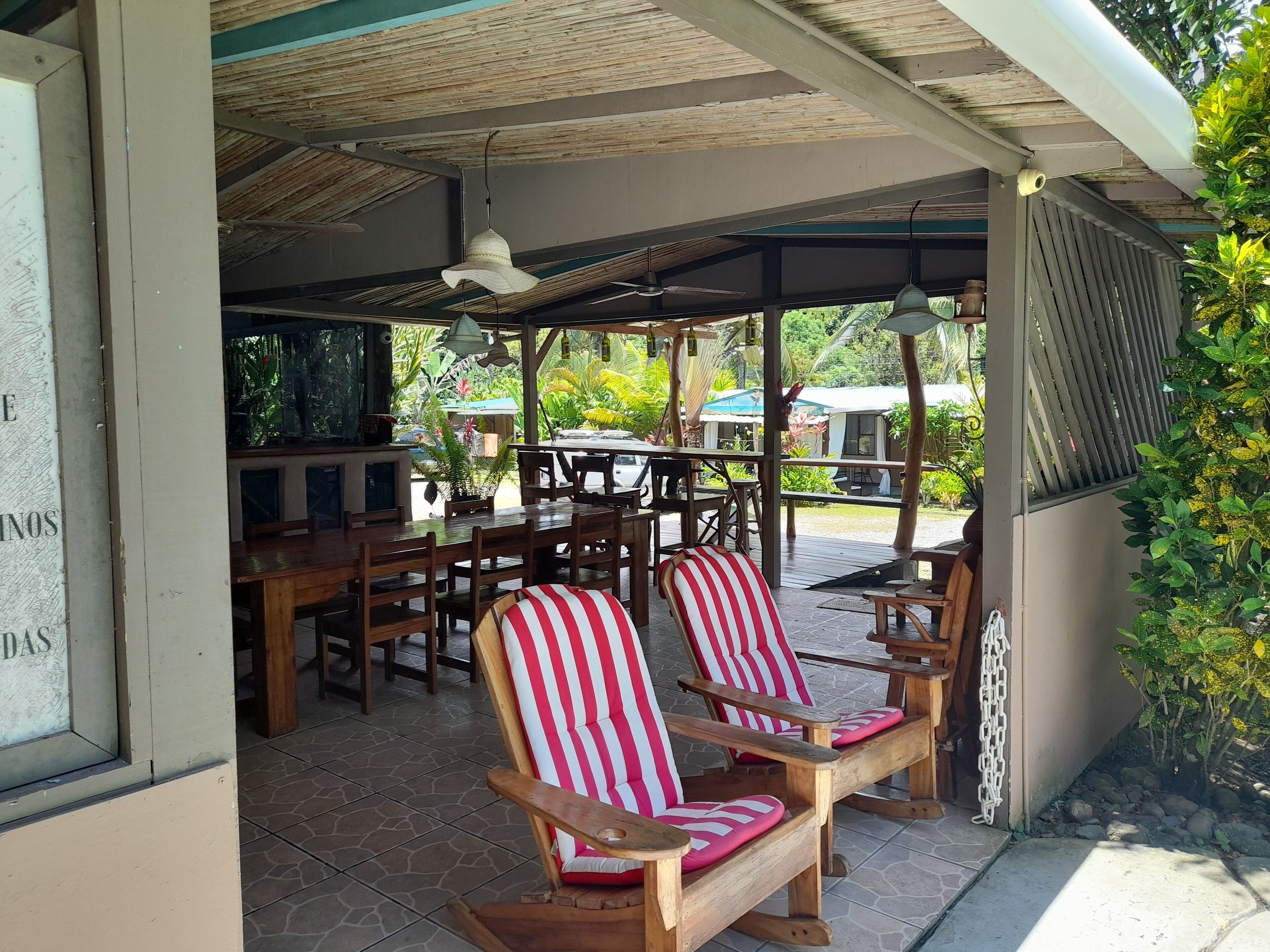 Un espace extérieur aménagé, abrité et doté de chaises et de tables en bois, entouré de verdure.
