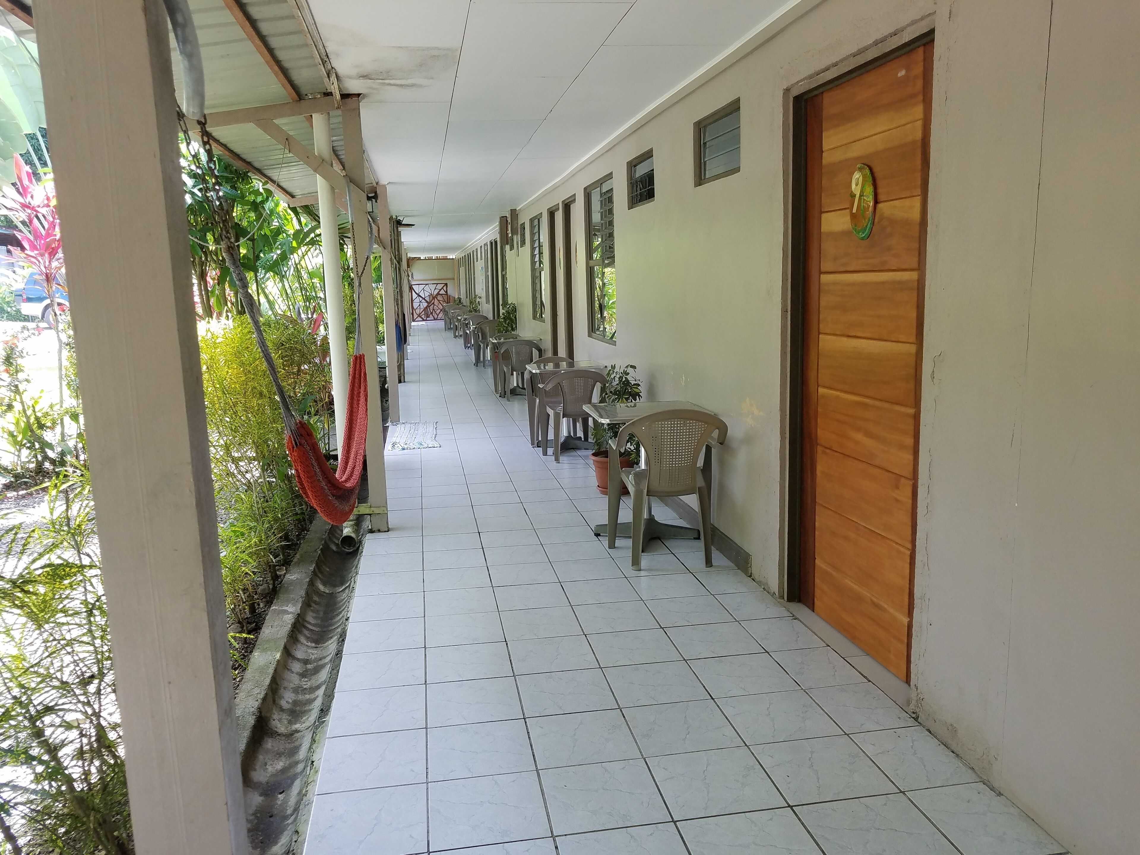 A corridor with tiled flooring, chairs, and tables arranged along the walls, with a door on the right and a hammock on the left.