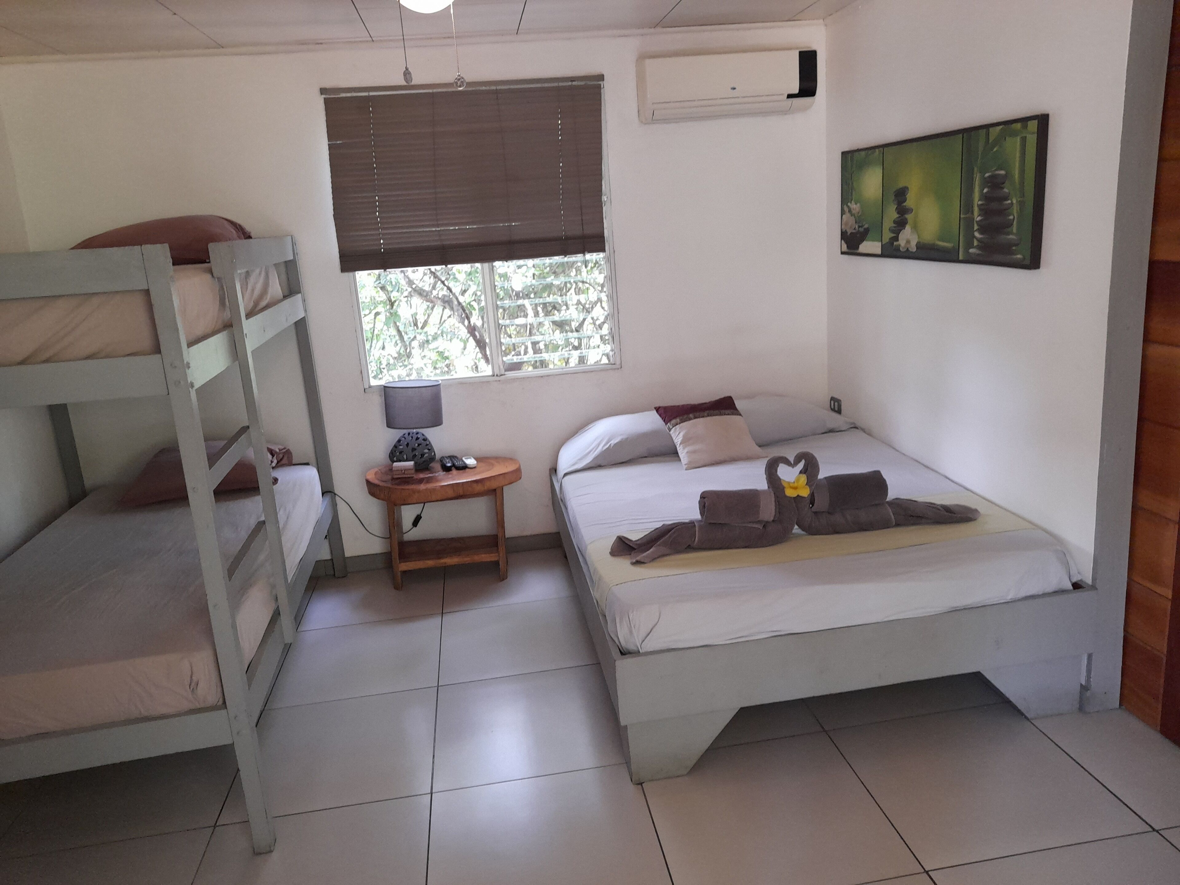 Un dormitorio con literas, una mesita de noche con lámpara, una ventana con persianas y un aire acondicionado de pared.