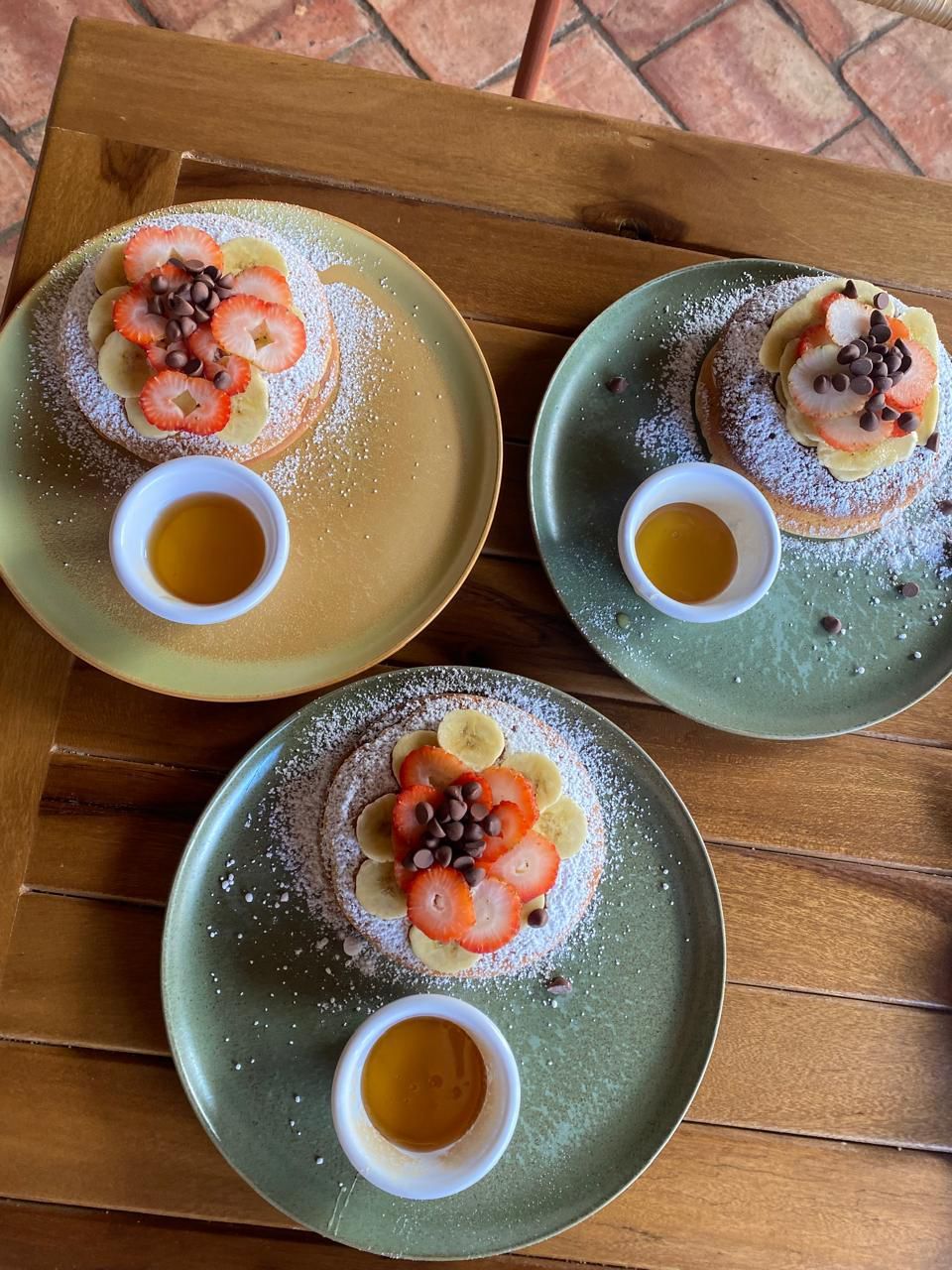 Drie borden met dessert, met aardbeien, bananen en chocoladestukjes, elk met een klein bakje siroop, op een houten tafel.