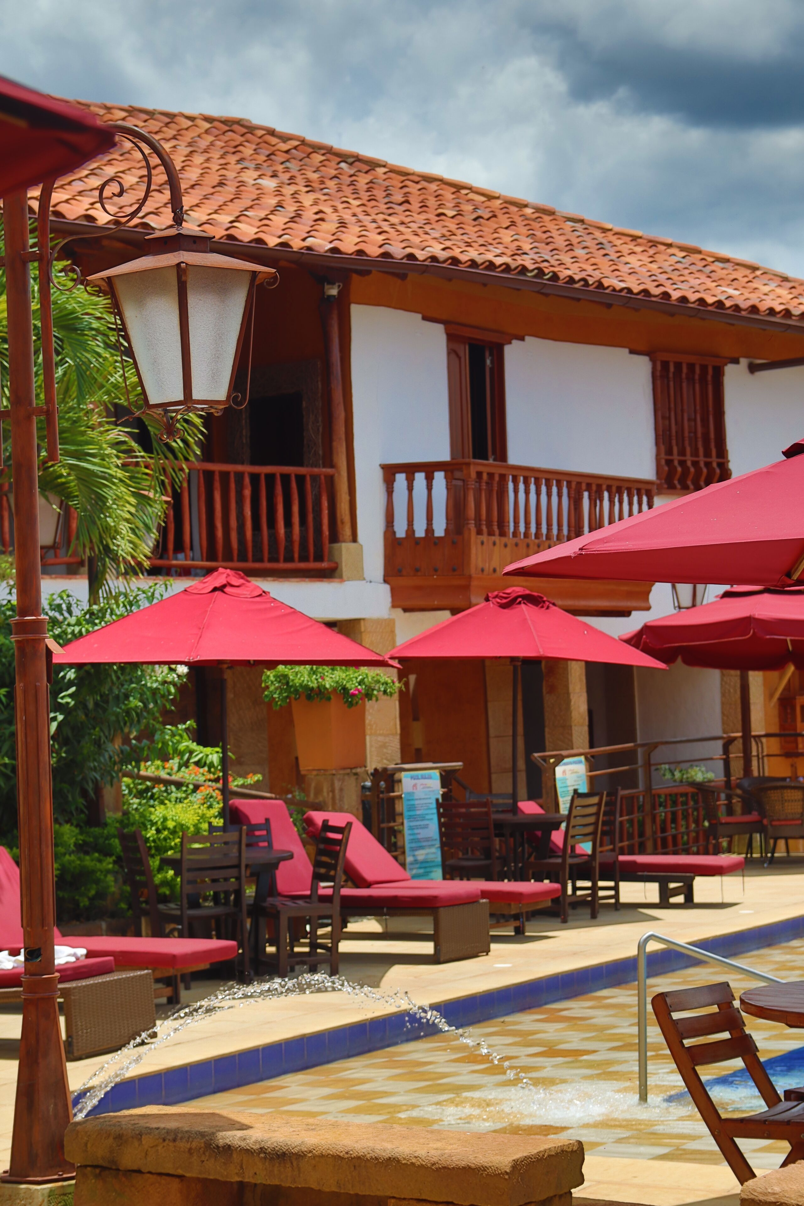 A poolside area with red umbrellas, lounge chairs, and a building with a tiled roof.