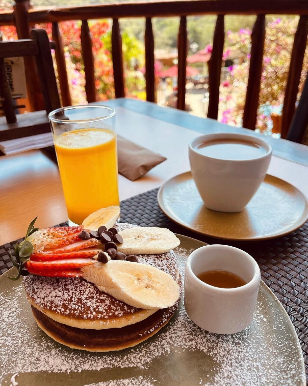 A breakfast setting with pancakes topped with strawberries, bananas, and chocolate chips, a glass of orange juice, a cup of coffee, and a cup of tea on a table with a view of flowers.
