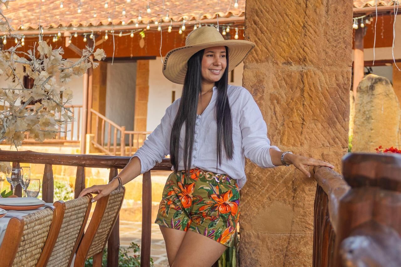 A person leaning on a wooden railing, wearing a hat and floral shorts, with a table and chairs in the foreground.