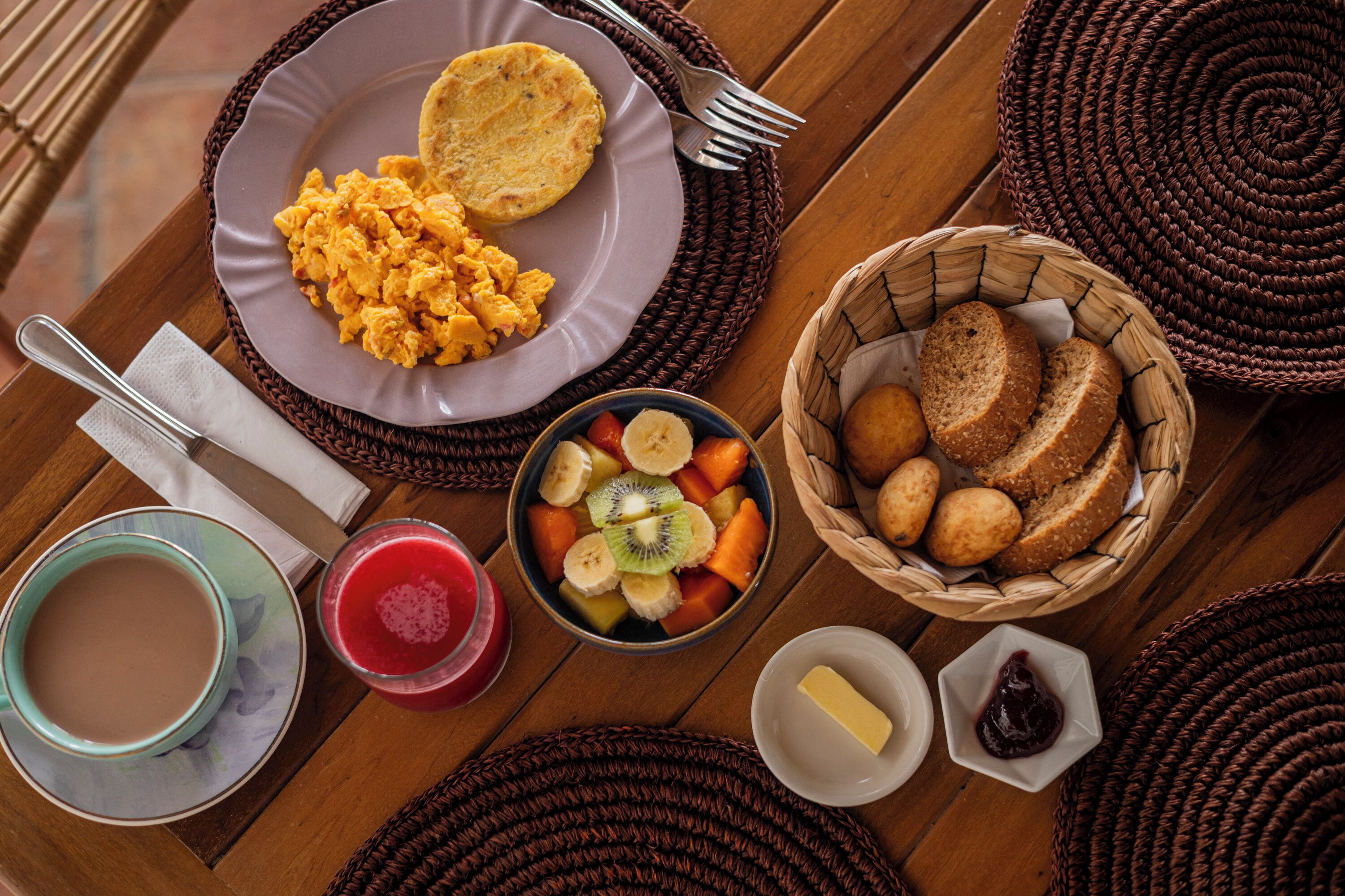 Une table dressée pour le petit-déjeuner avec des œufs brouillés, des toasts, un bol de fruits, des petits pains, une tasse de café, un verre de jus de fruits, un pot de beurre et un pot de confiture.
