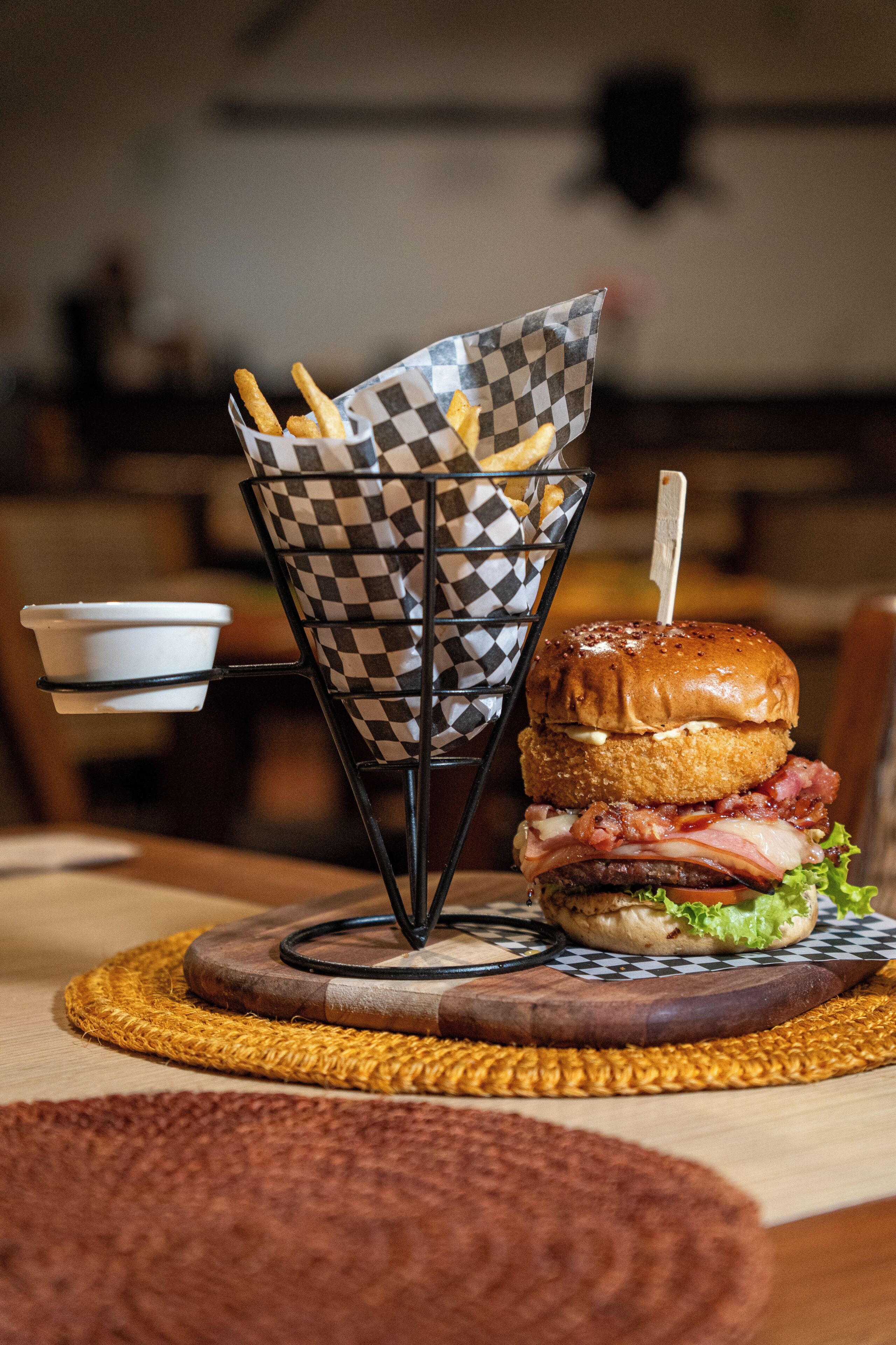 A burger with fries on a checkered paper basket, a cup, and a wooden table with a woven placemat.