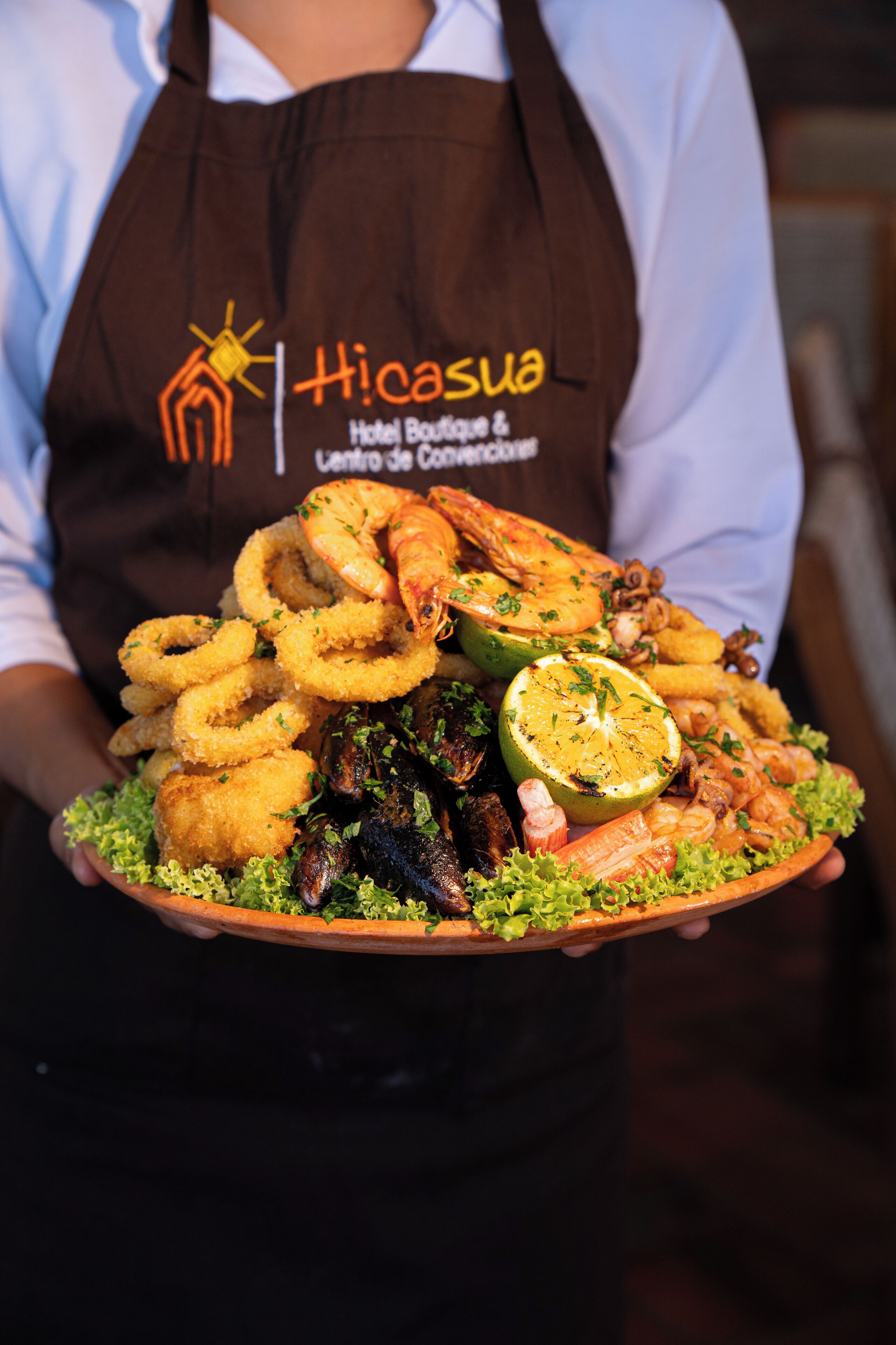 A person holding a platter with various seafood items, including shrimp, squid rings, and a lemon wedge.