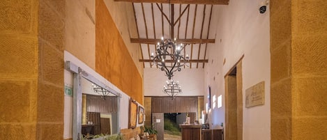A hallway with a chandelier, wooden beams, stone walls, a reception desk, and wicker furniture.