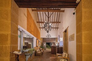 A hallway with a chandelier, wooden beams, stone walls, a reception desk, and wicker furniture.