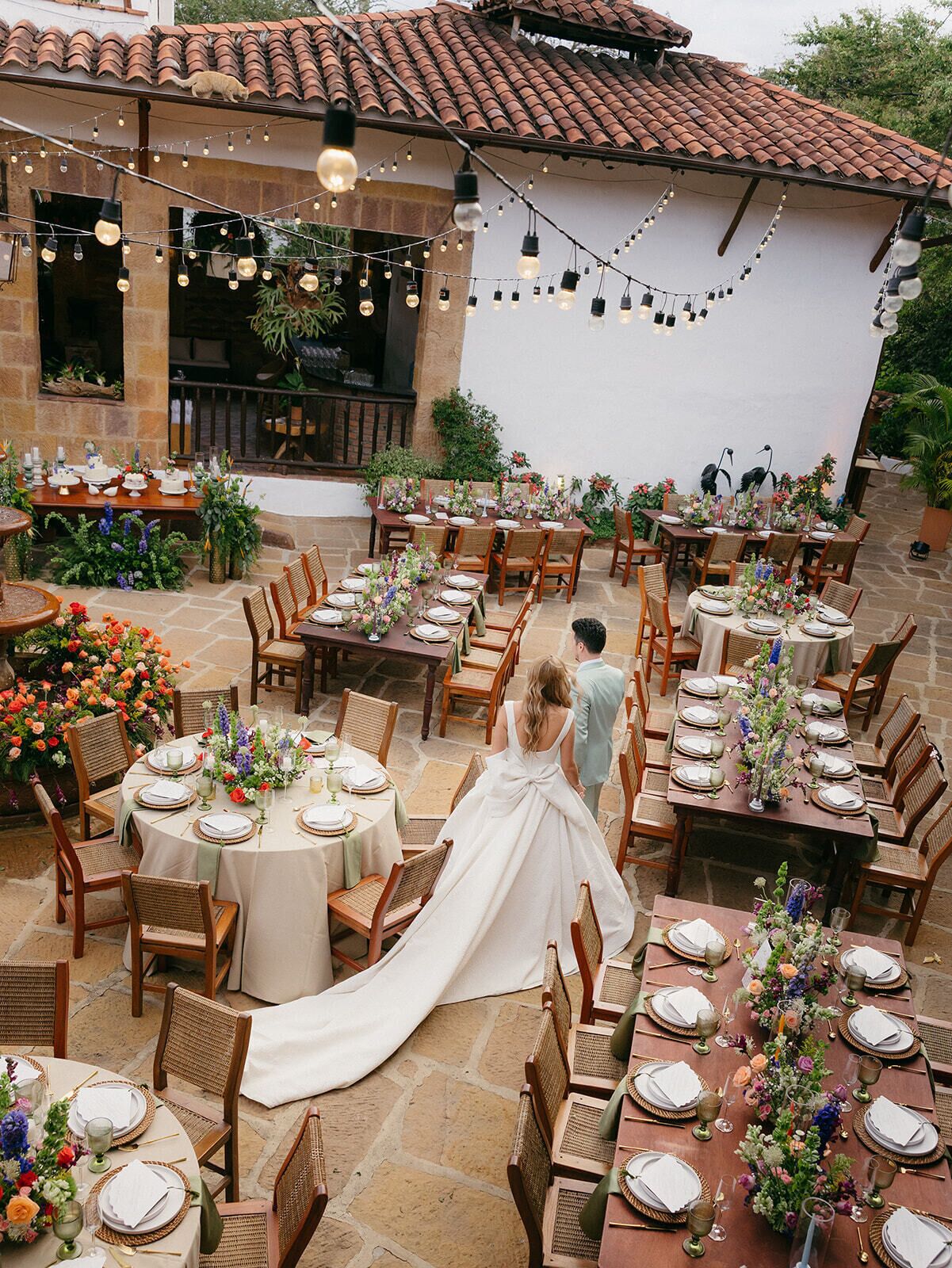 Un salón de bodas al aire libre con varias mesas redondas, sillas e iluminación con luces colgantes.