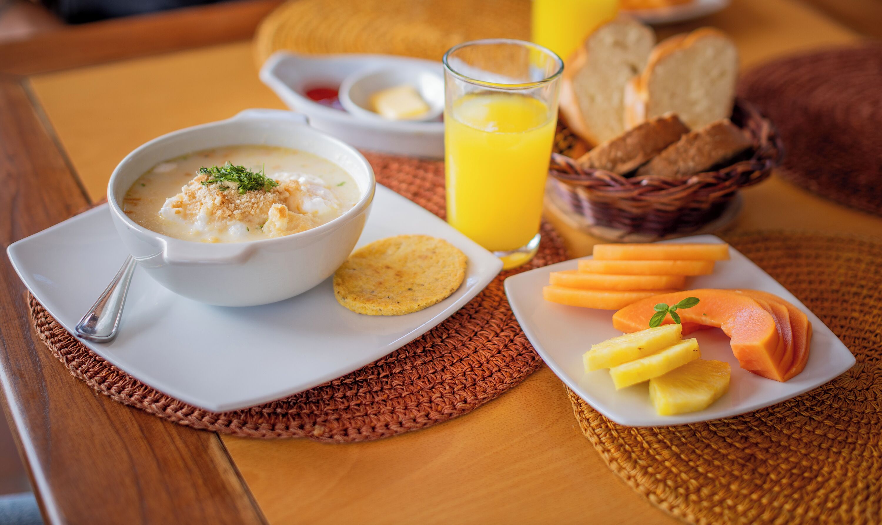 Uma mesa de café da manhã com uma tigela de sopa, torradas, um copo de suco de laranja e um prato com frutas e biscoitos.