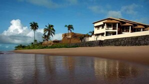 A beachfront property with a two-story house, a stone wall, and palm trees. - Pousada Restinga (Lauro de Freitas)