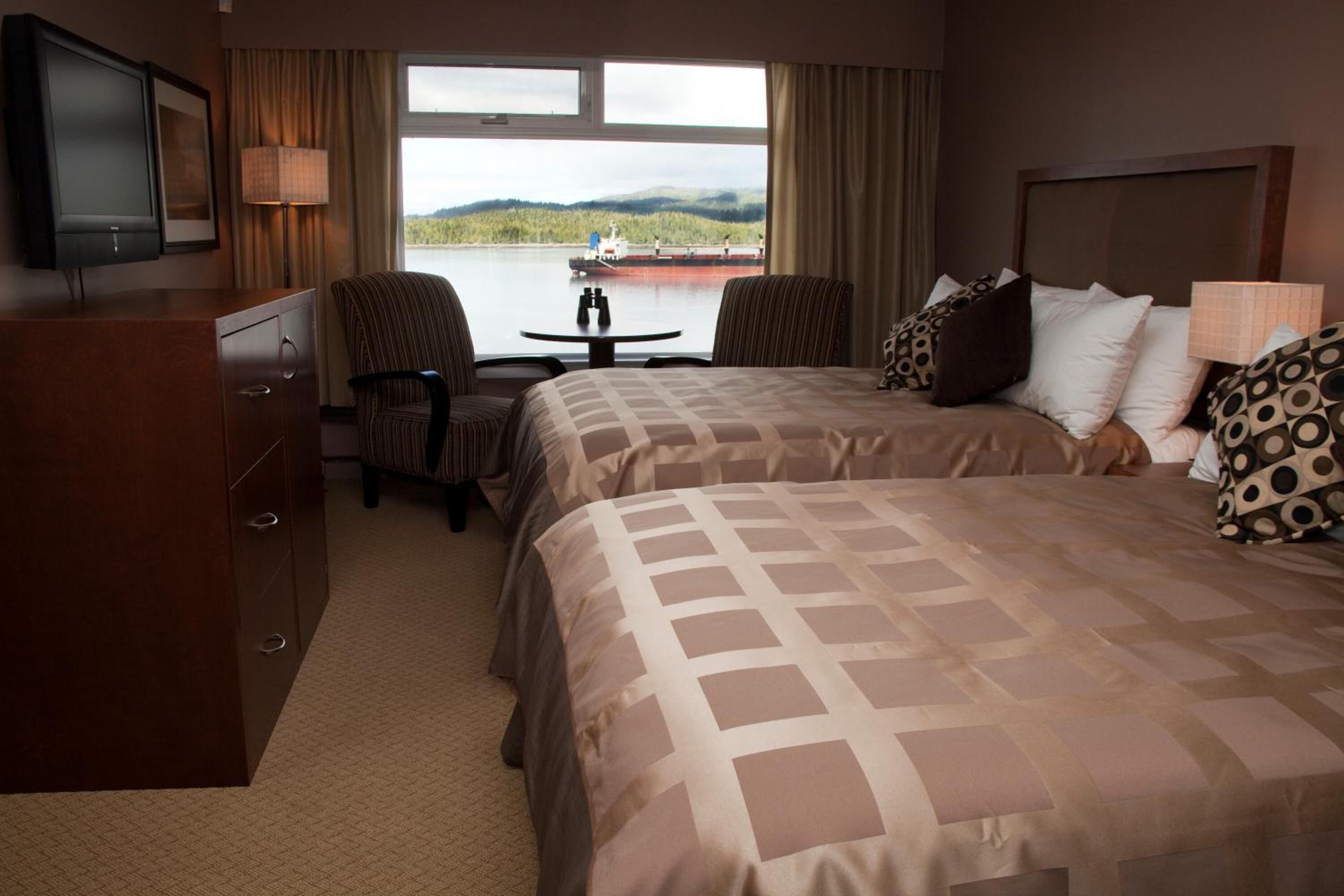 A hotel room with two beds, a desk, a chair, a TV, and a view of a lake with a boat.