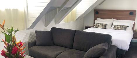 A modern bedroom with a dark grey sofa, a glass coffee table, and a bed with white bedding and patterned pillows.