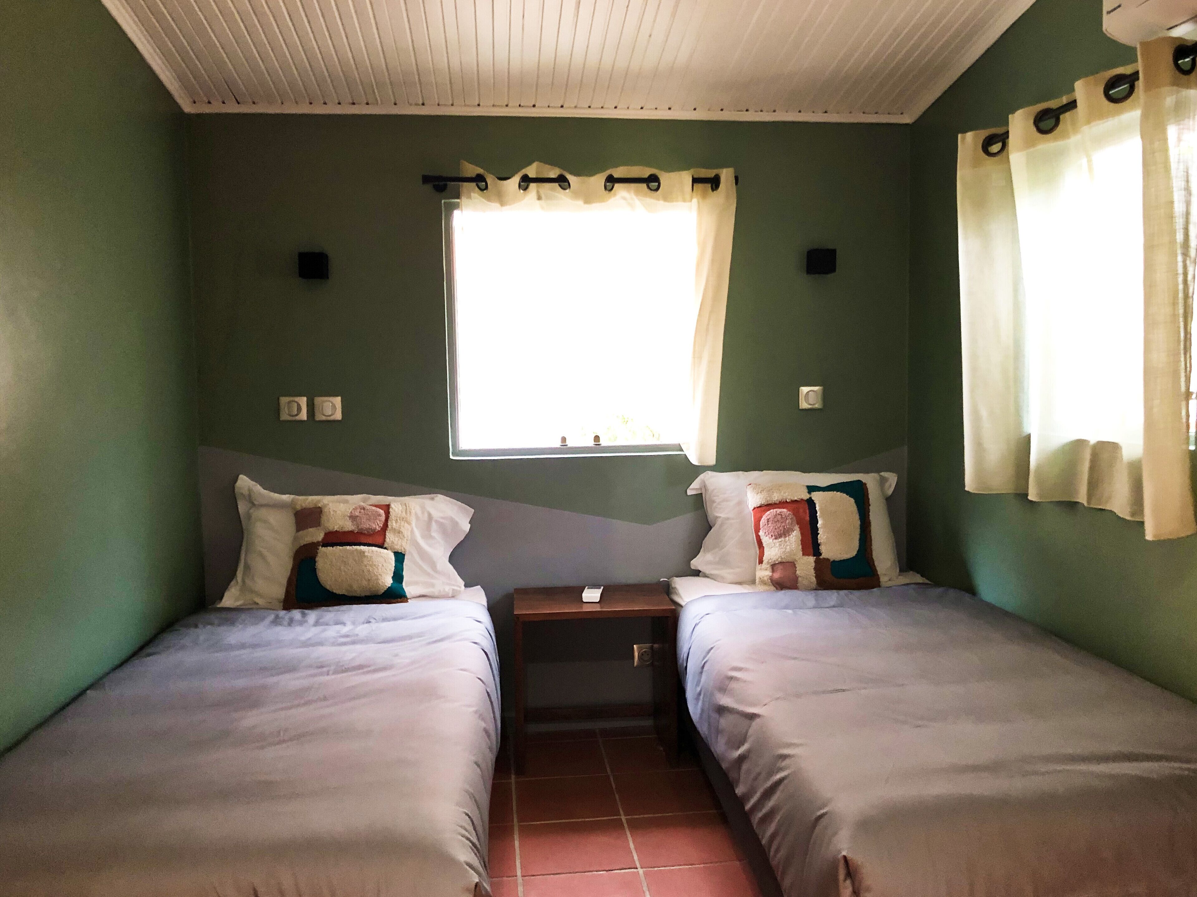 A small, single-room accommodation with two single beds, a window with curtains, and a tiled floor.