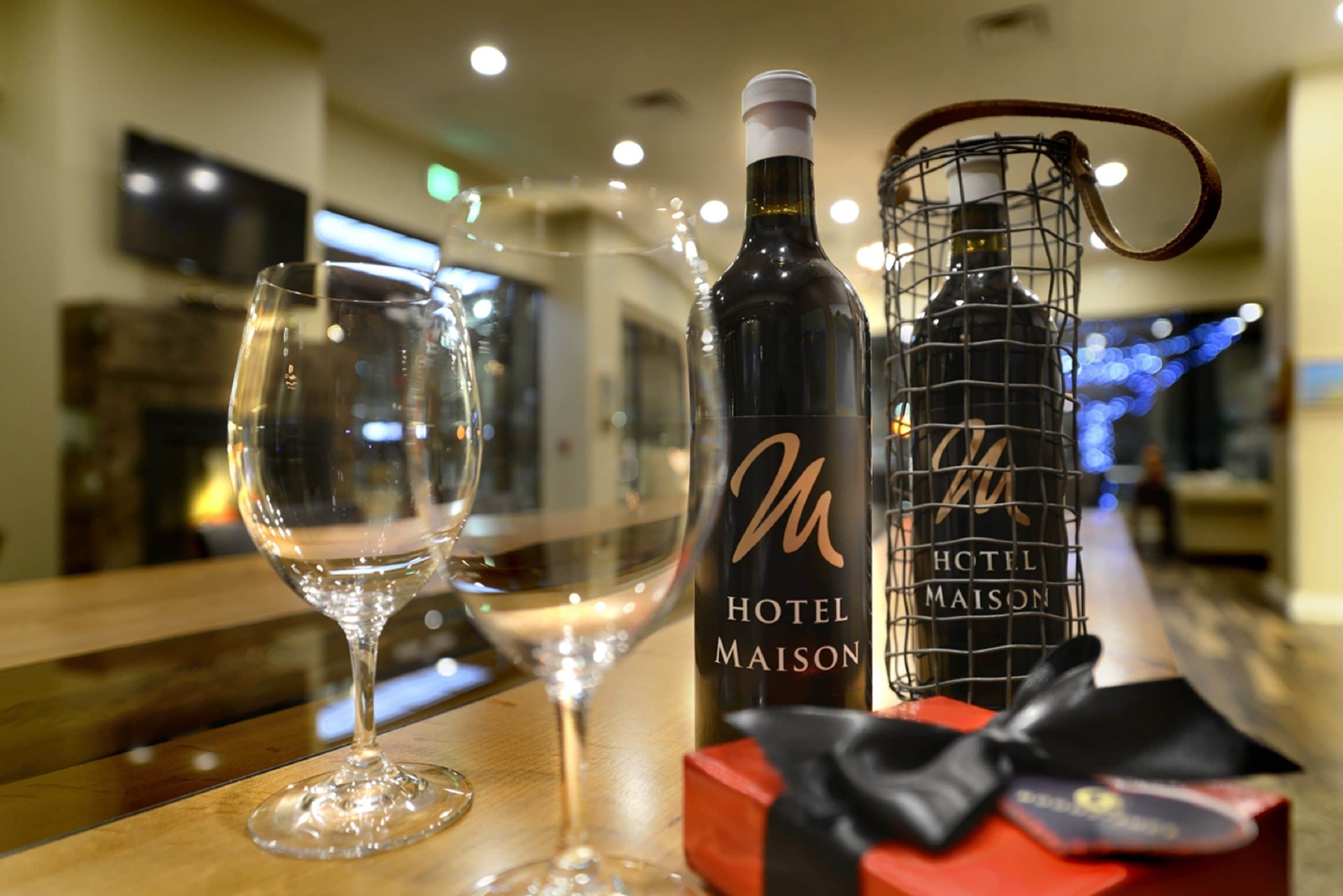 A bottle of Hotel Maison wine, a wine glass, and a gift box on a wooden table in a hotel setting.