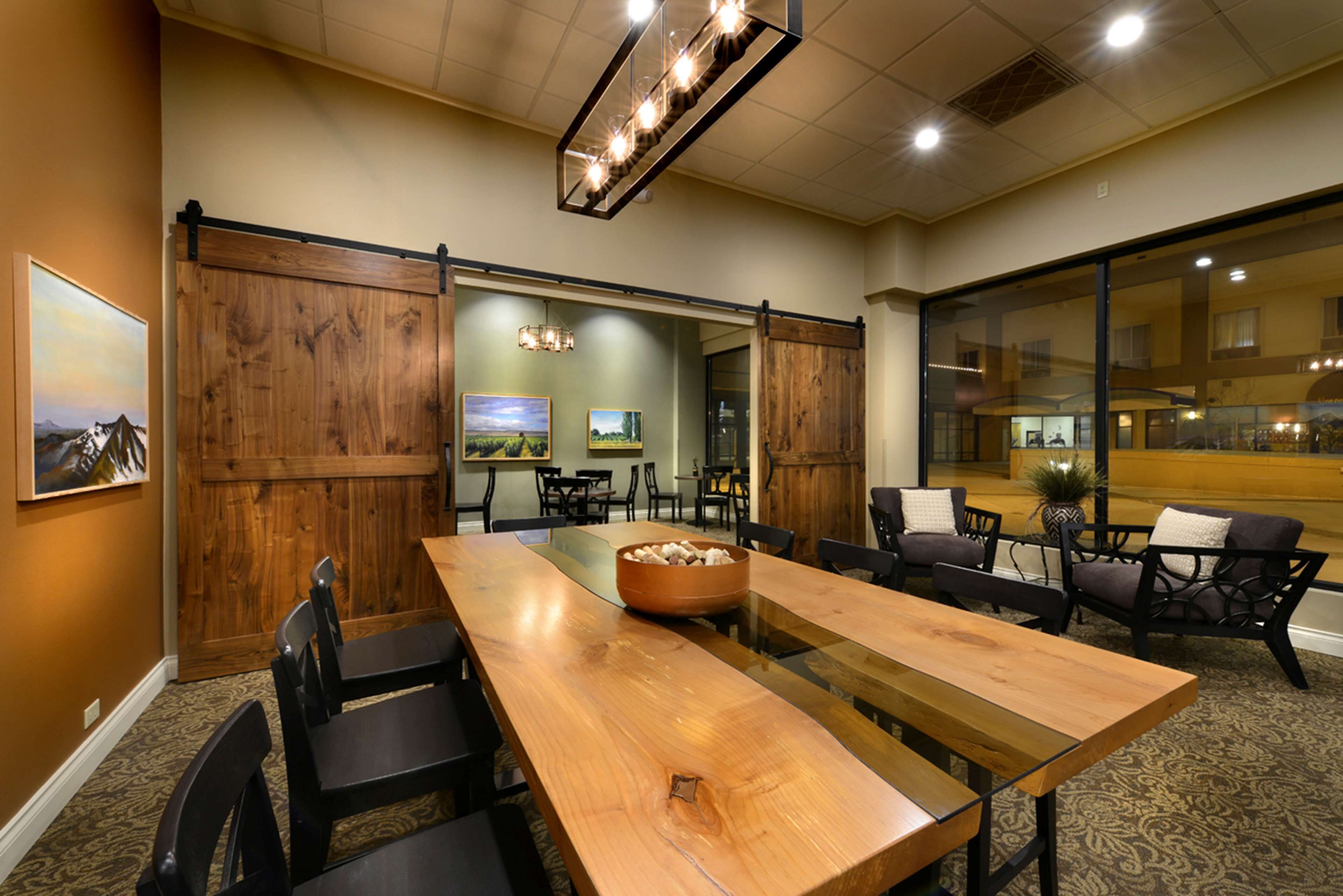 A conference room with a long wooden table, black chairs, and a glass partition. There are framed pictures on the walls and a chandelier above the table.