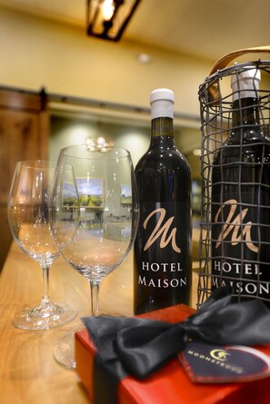A table with two wine glasses, two bottles of Hotel Maison wine, a metal wine rack, and a red box with a black bow.